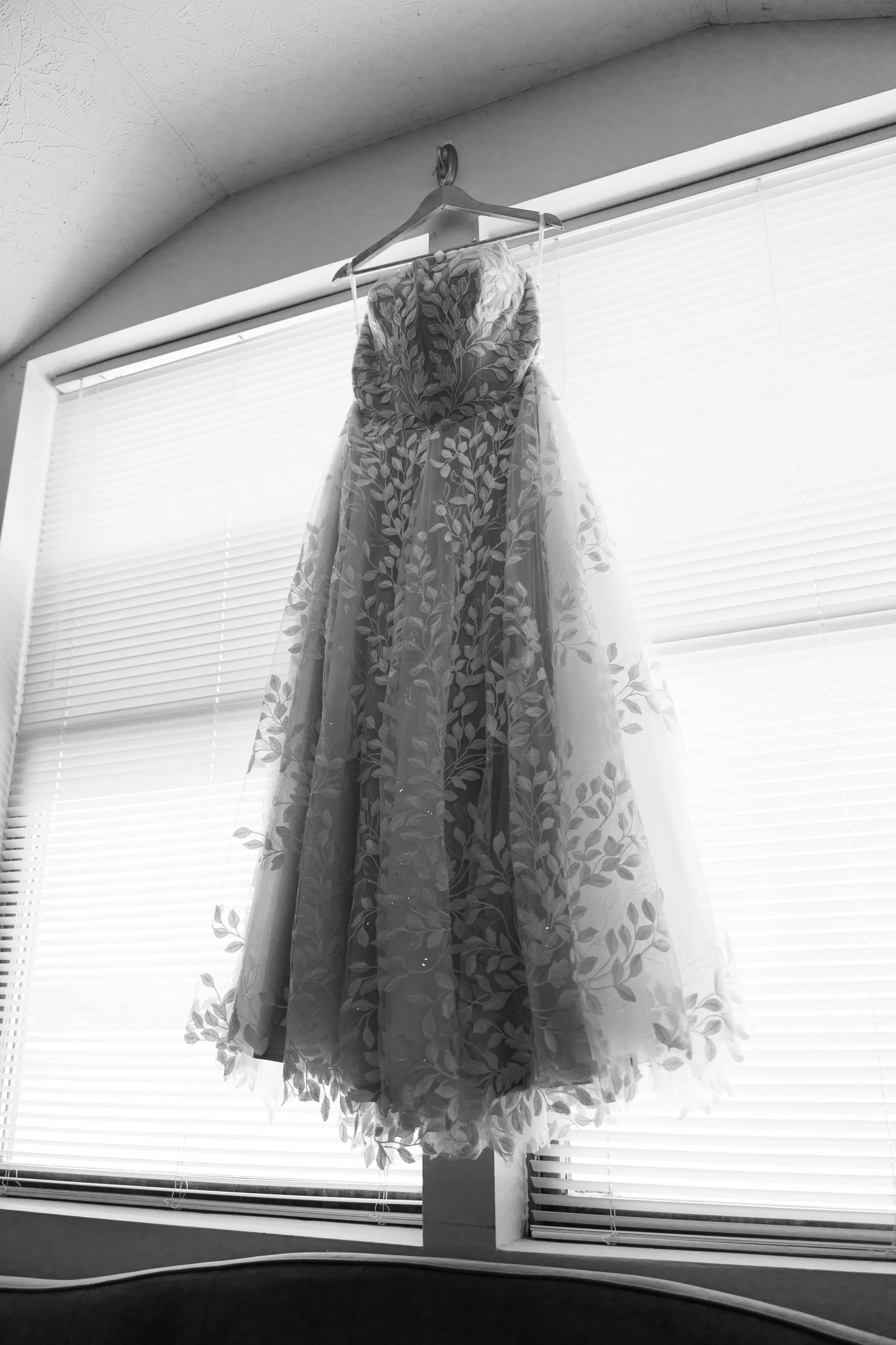 A wedding dress hanging on a hanger in front of a window with blinds.