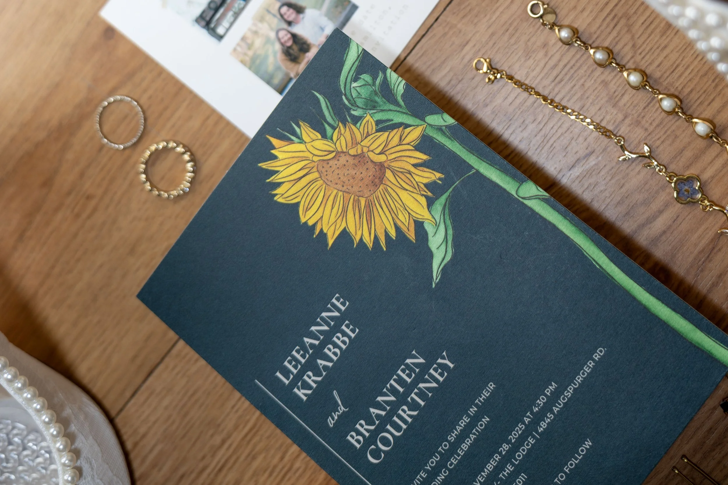 A wedding invitation for Leanne Krabbe and Brantyn Courtney with a sunflower design, placed on a wooden surface with pearl jewelry, gold rings, and a pearl necklace