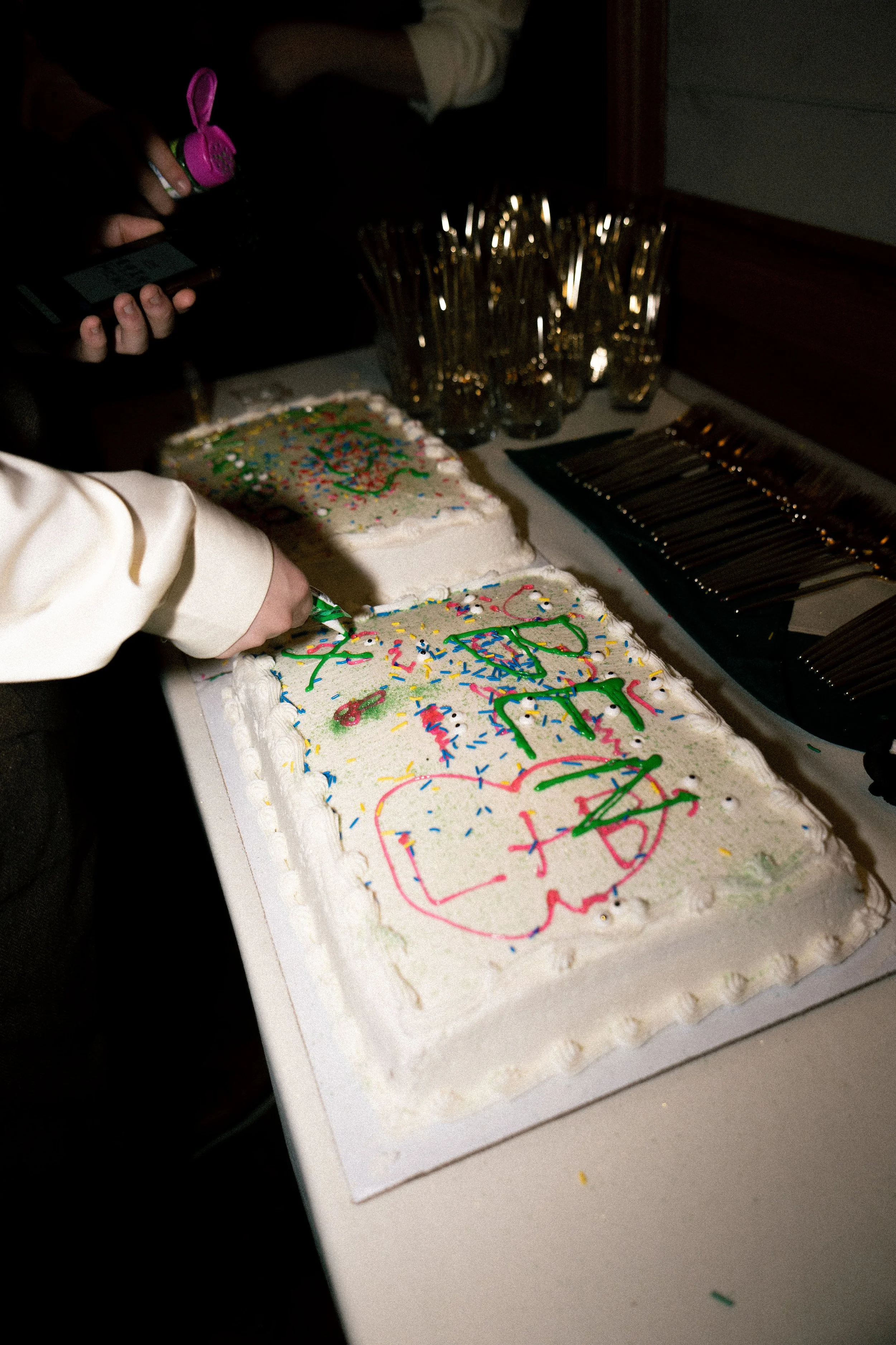 Person cutting a birthday cake with colorful sprinkles and frosting decorations, surrounded by glasses and utensils at a party.