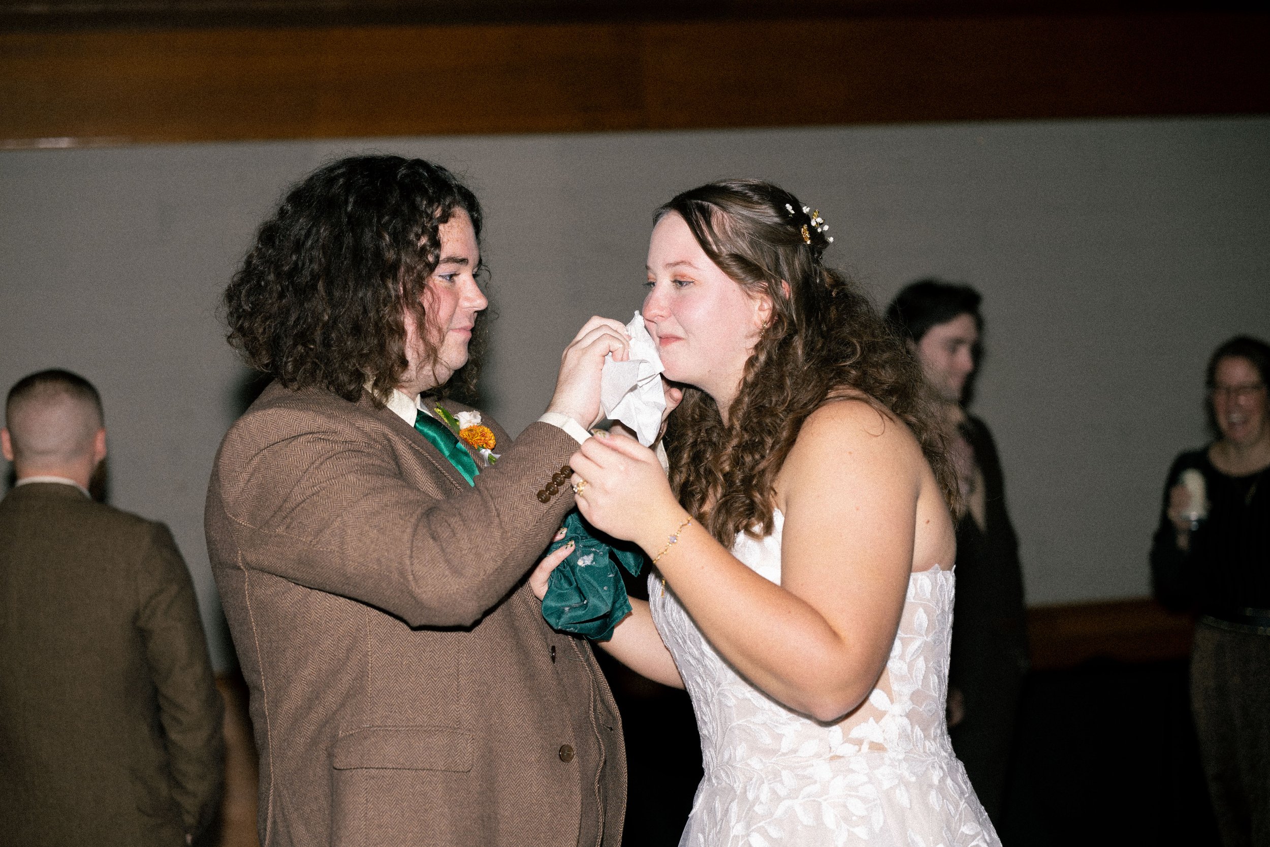 A bride with curly hair wearing a white wedding dress is crying while wiping her tears with tissue, being comforted by a person in a brown suit.