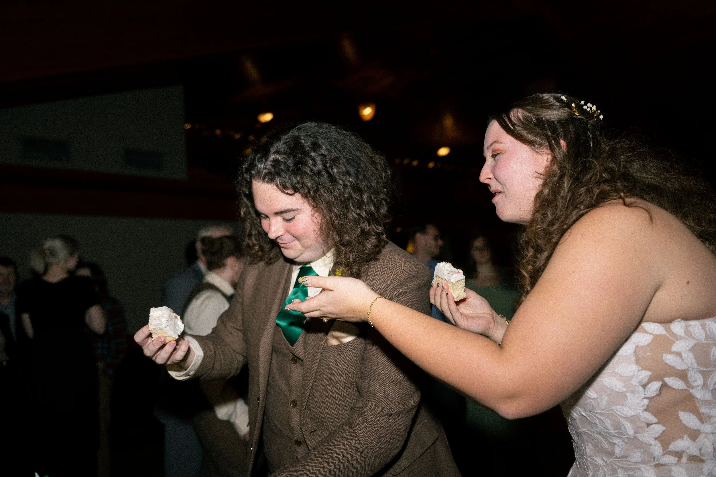 A woman feeding a man a piece of cake at a wedding reception, with other guests in the background.
