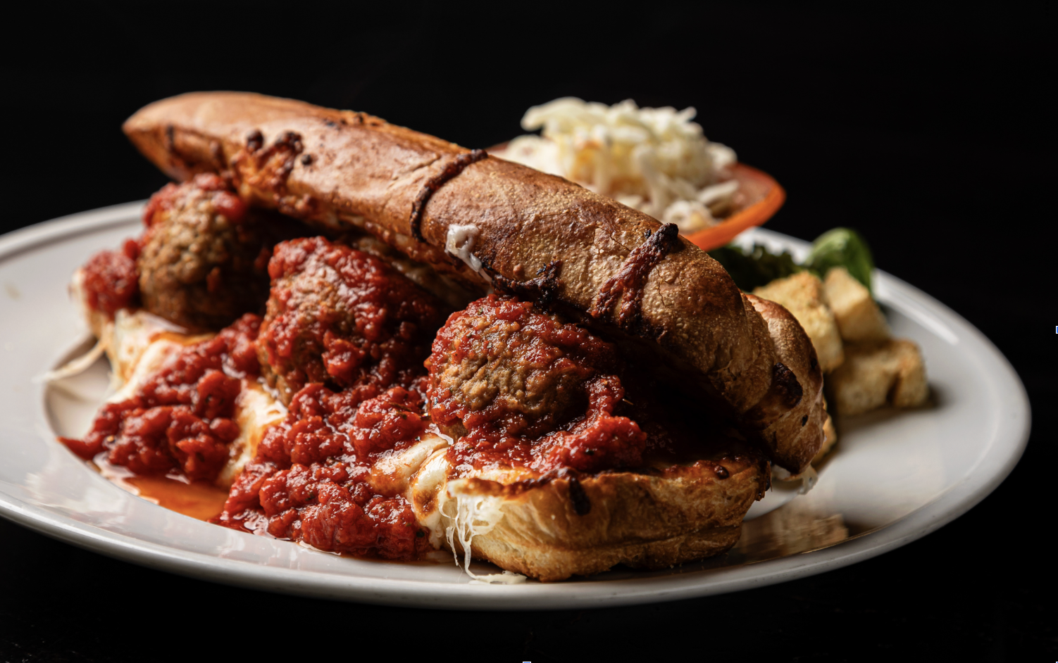 Plate of Italian spaghetti with meatballs, topped with marinara sauce, served with garlic bread, pasta salad, and croutons against a black background.