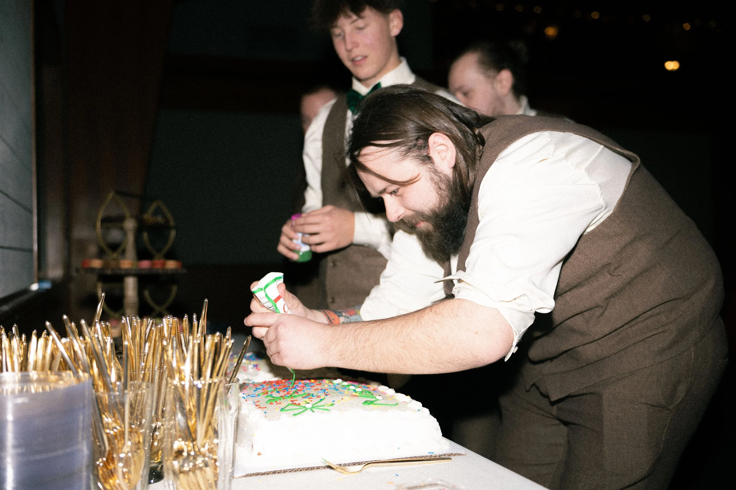 A man with a beard and long dark hair is leaning over a table, decorating a white cake with colorful sprinkles. Two young boys in formal attire stand behind him, holding small bottles of frosting or decorations, at a festive event.
