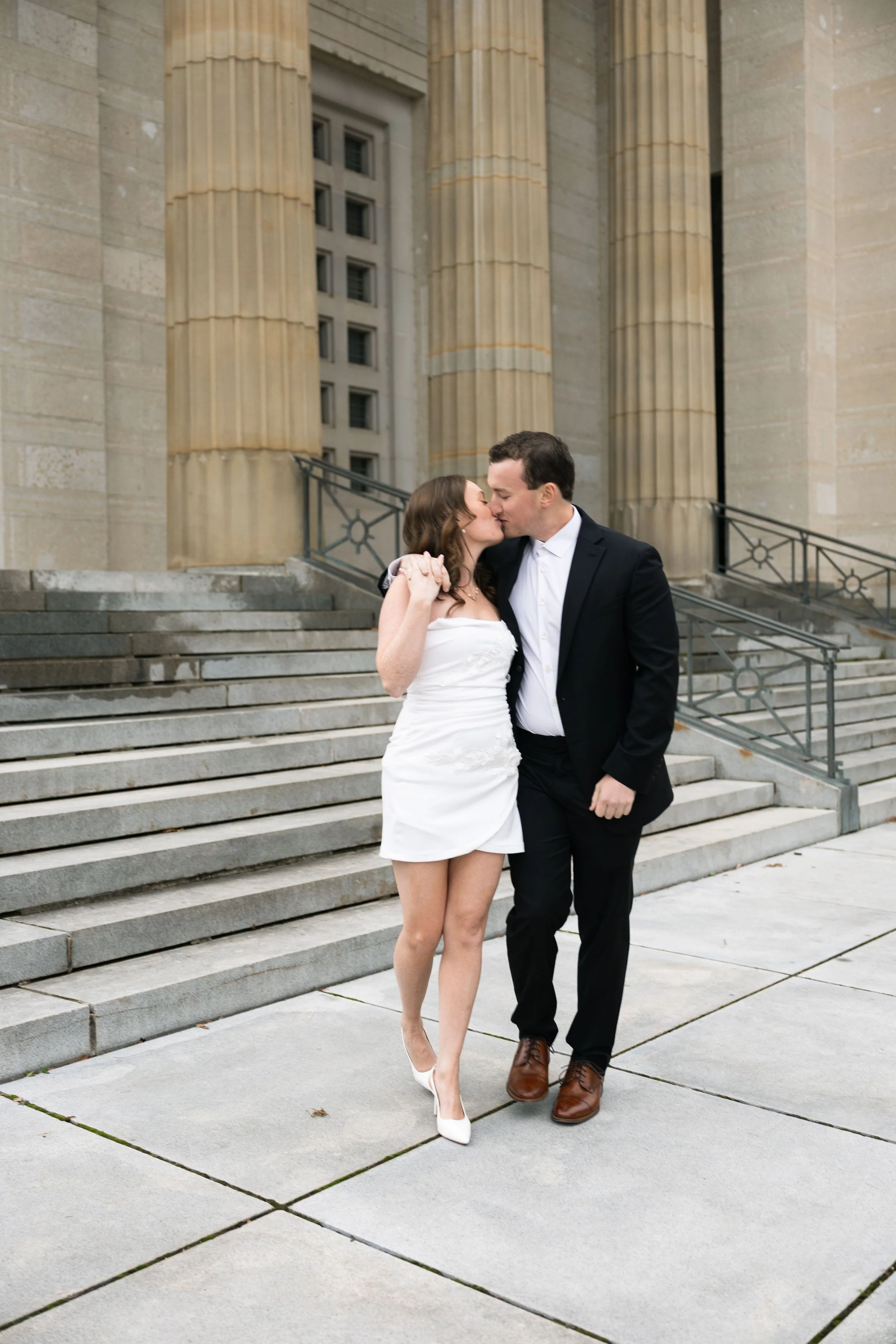 A couple dressed formally, the woman in a white dress and heels, and the man in a dark suit and brown shoes, standing on steps outside a historic building with large columns, sharing a kiss.
