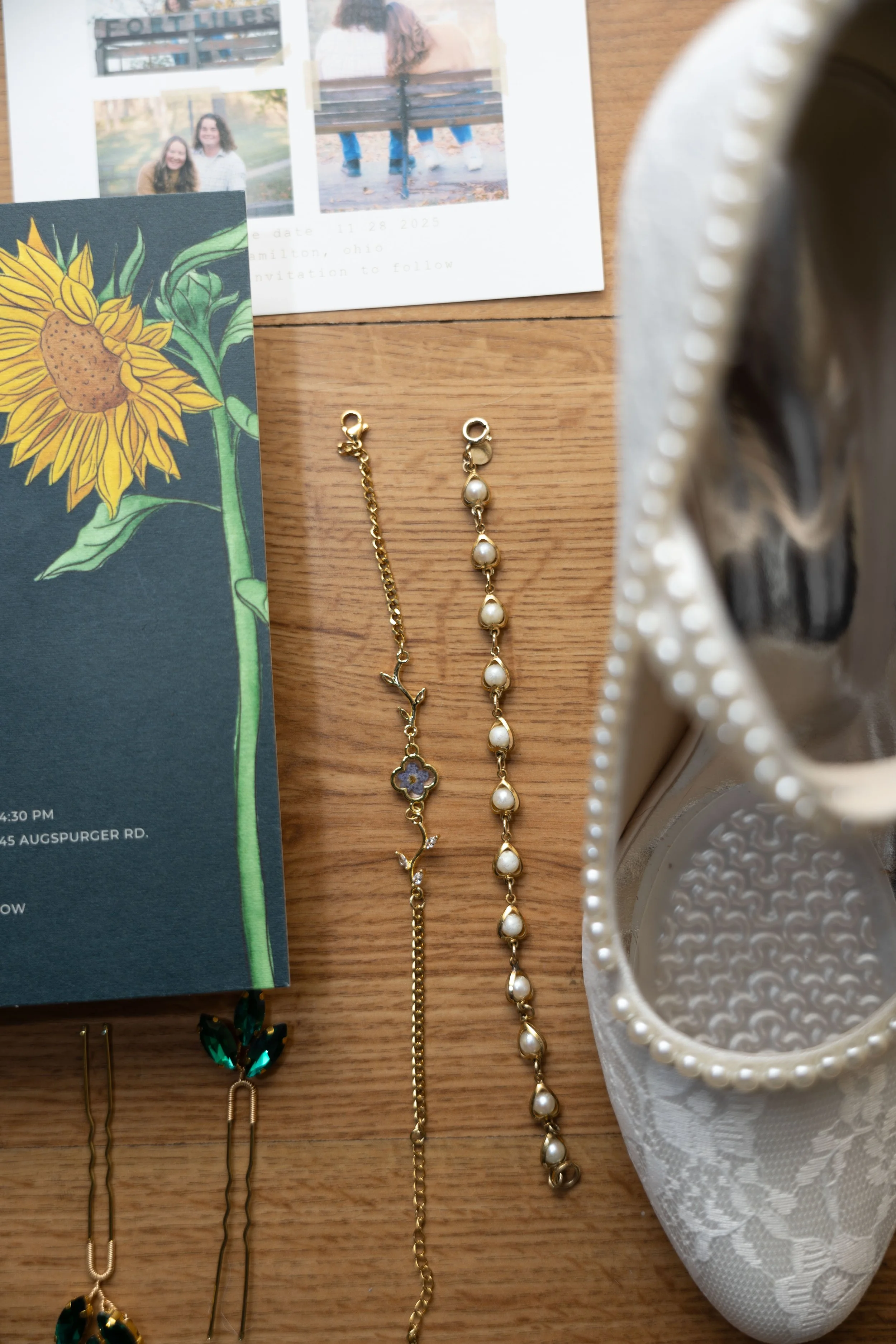 A wooden table with a floral brochure, a photo collage, two gold bracelets with charms, pearl earrings, a white lace shoe with pearl embellishments, and a white ceramic cup.