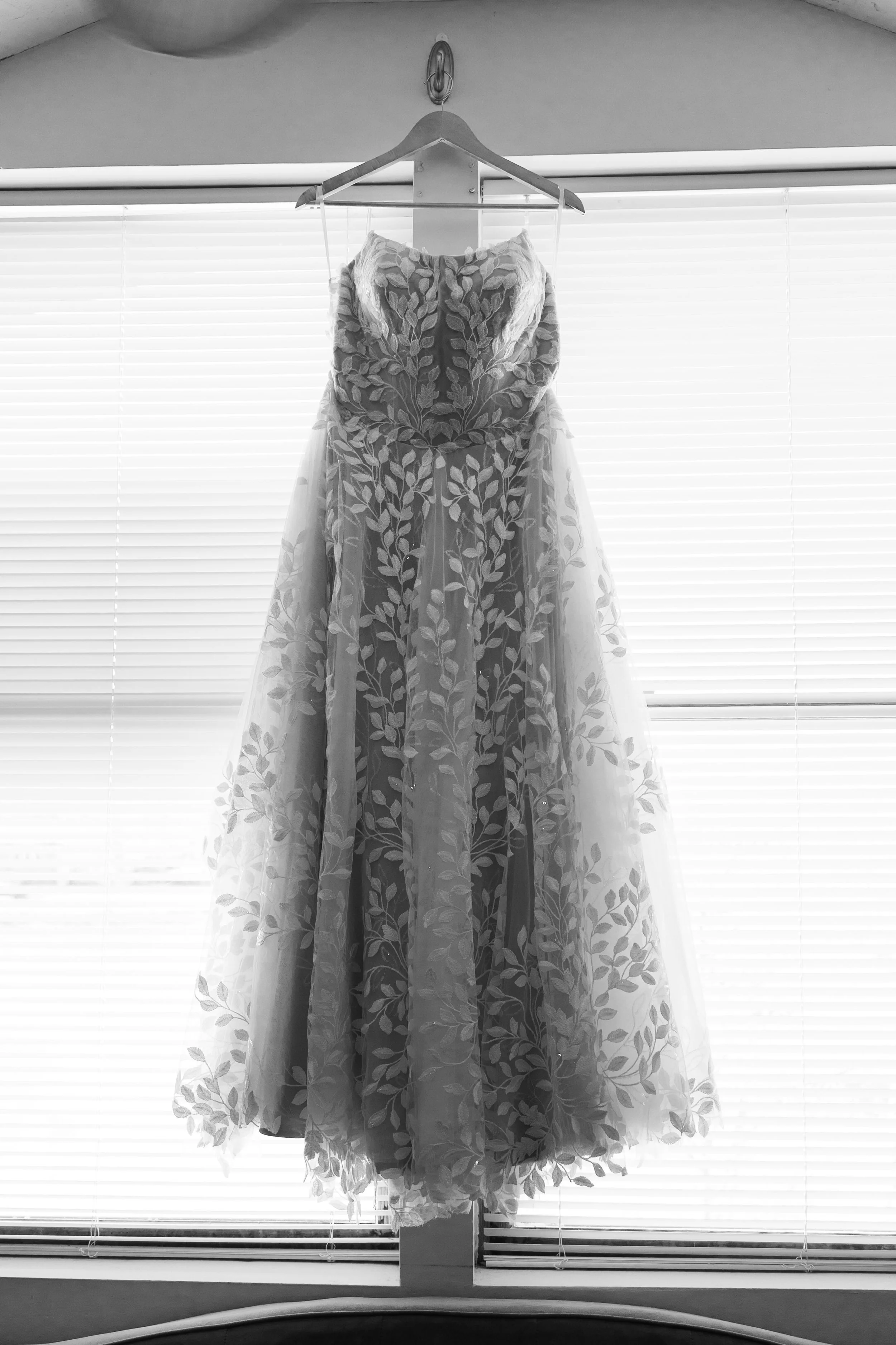 A wedding dress hanging on a hanger in front of a window with blinds.