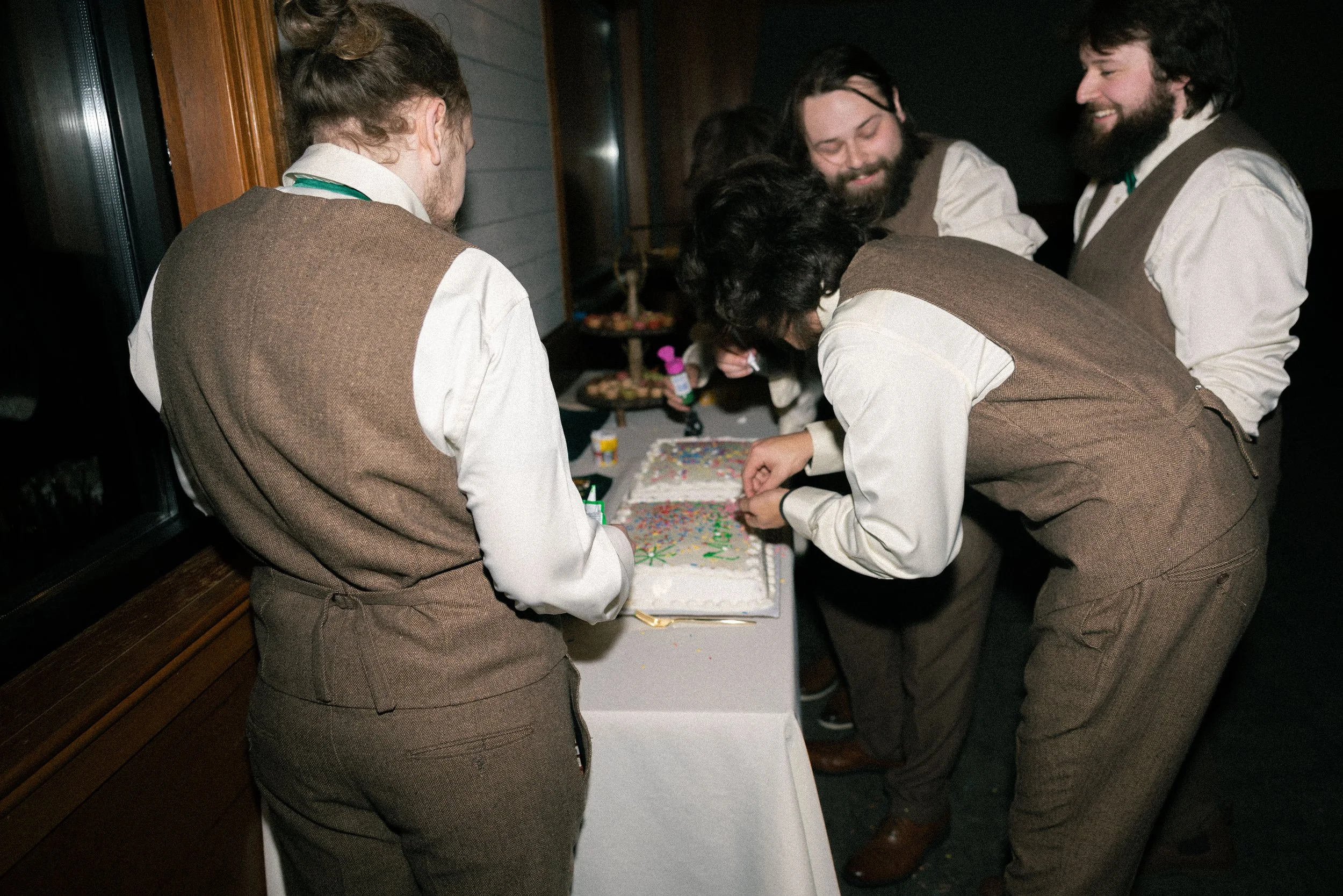 Group of people in vintage-style clothing gathered around a decorated birthday cake, celebrating and preparing to cut it.