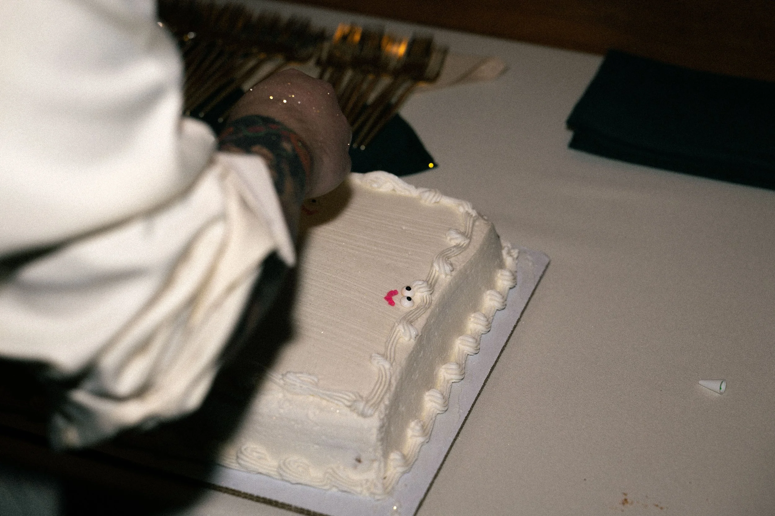A white rectangular cake with piped borders and a small face with eyes and a pink heart on top, on a table with a person's arm and hand, partially visible, reaching for the cake.