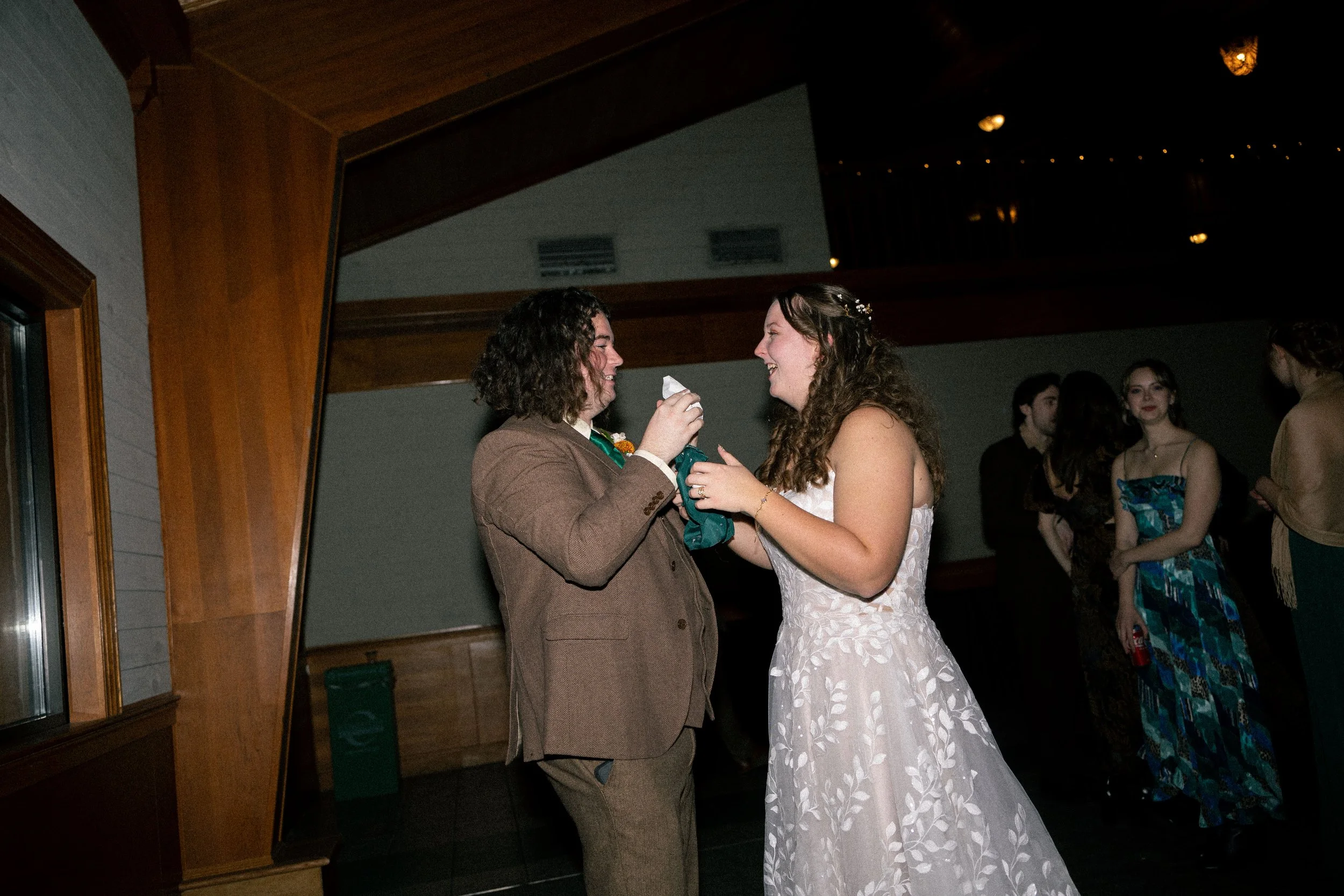 A bride and groom sharing a moment at their wedding reception, surrounded by guests, with the bride in a white dress and the groom in a brown suit.