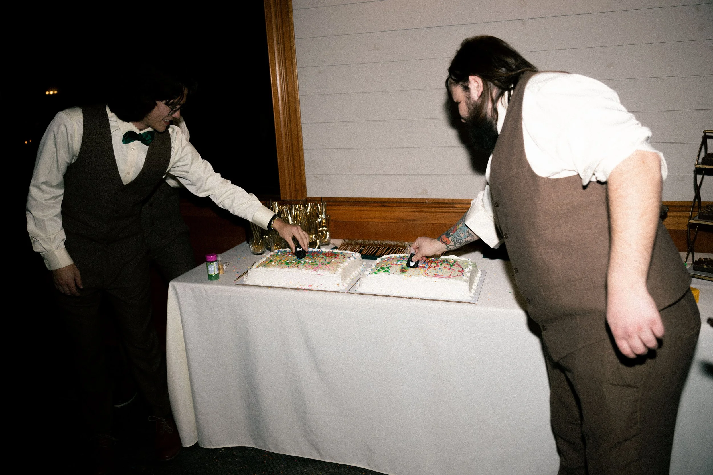 Two men with long hair and beards, dressed in vintage-style vests and white shirts, are cutting cakes at a celebration event.