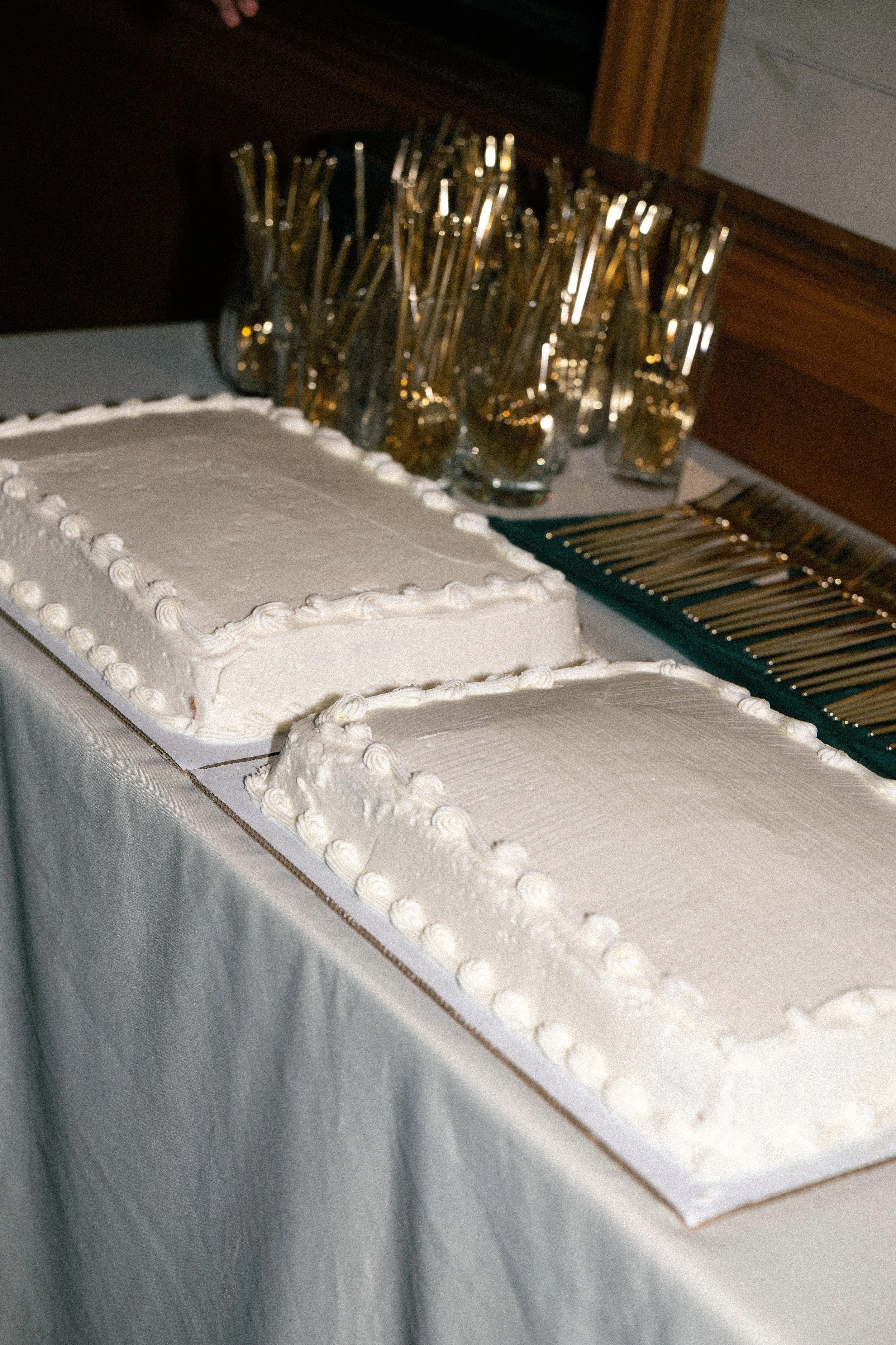 Two rectangular white frosted cakes on a table with gold-and-silver utensils in the background.