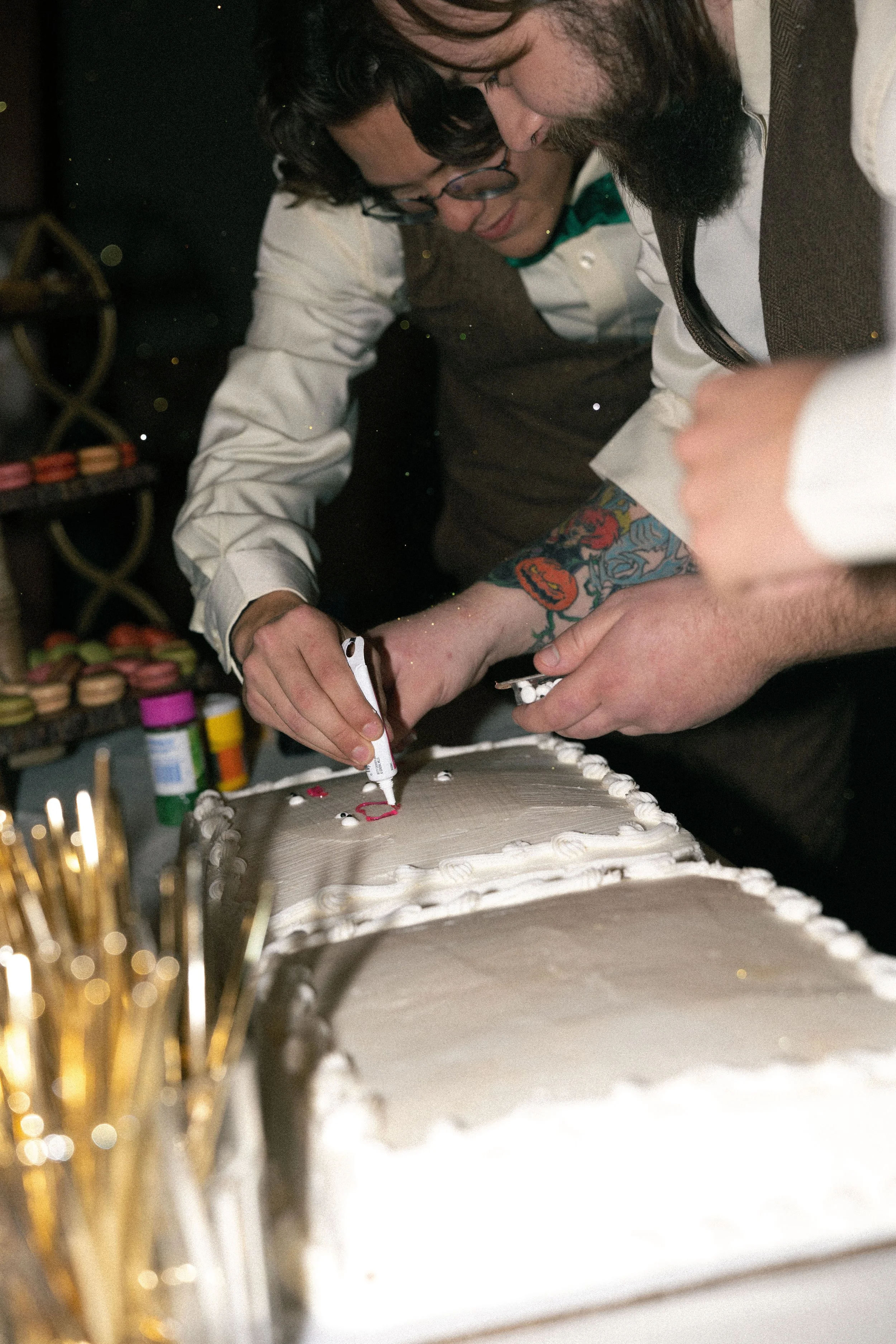 Two people are decorating a wedding cake with white icing, using markers to write on it.