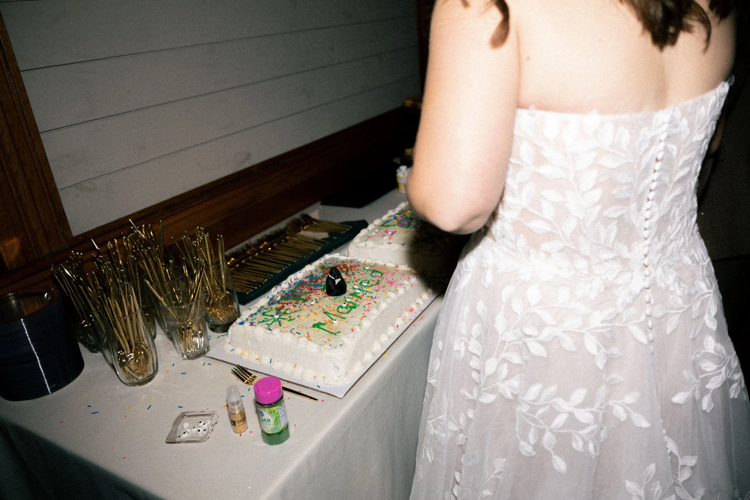 A woman in a white, floral-patterned dress stands near a table with a birthday cake decorated with colorful sprinkles and a small black figure on top, surrounded by empty glasses and birthday supplies.