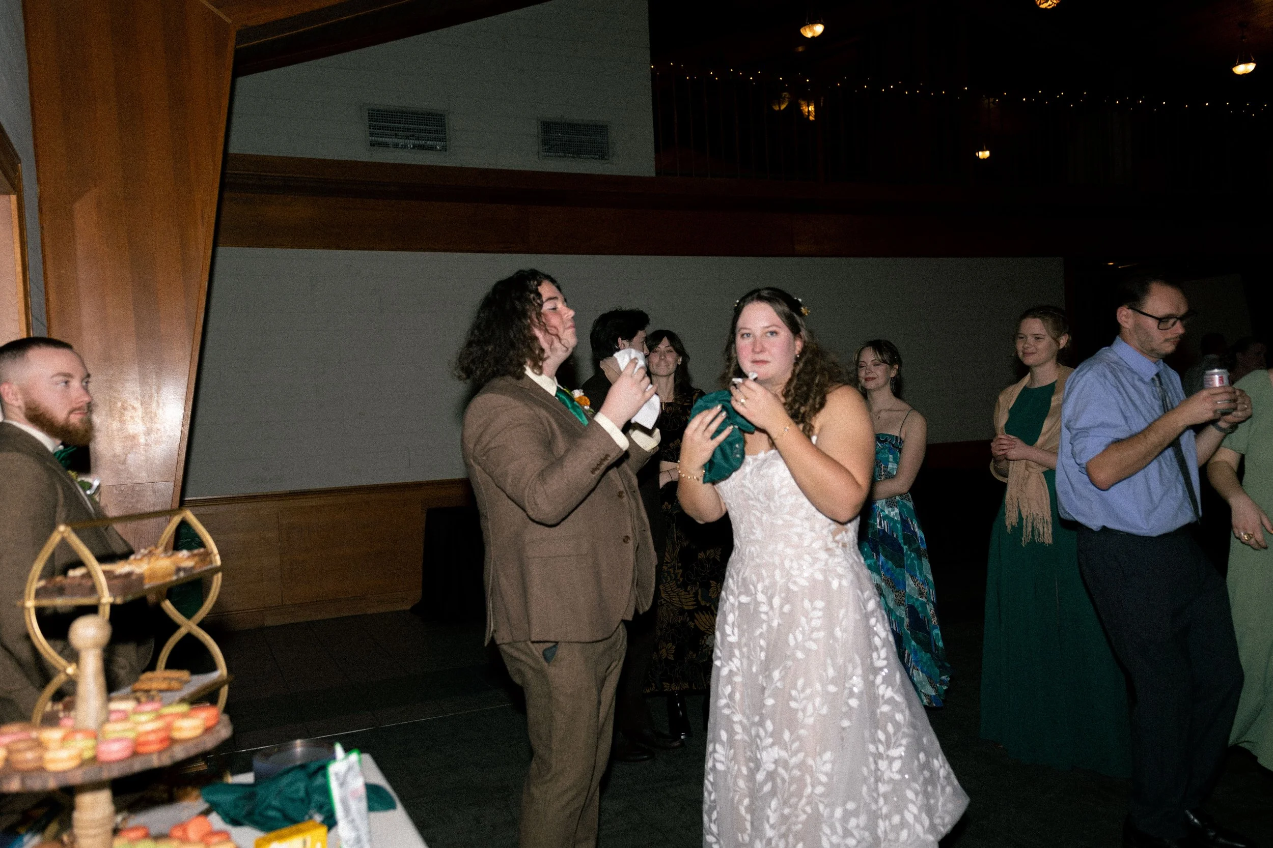 A wedding reception with guests, including a woman in a white wedding dress and a man in a brown suit, standing together and eating cake. Other guests are in the background, some holding drinks and chatting.