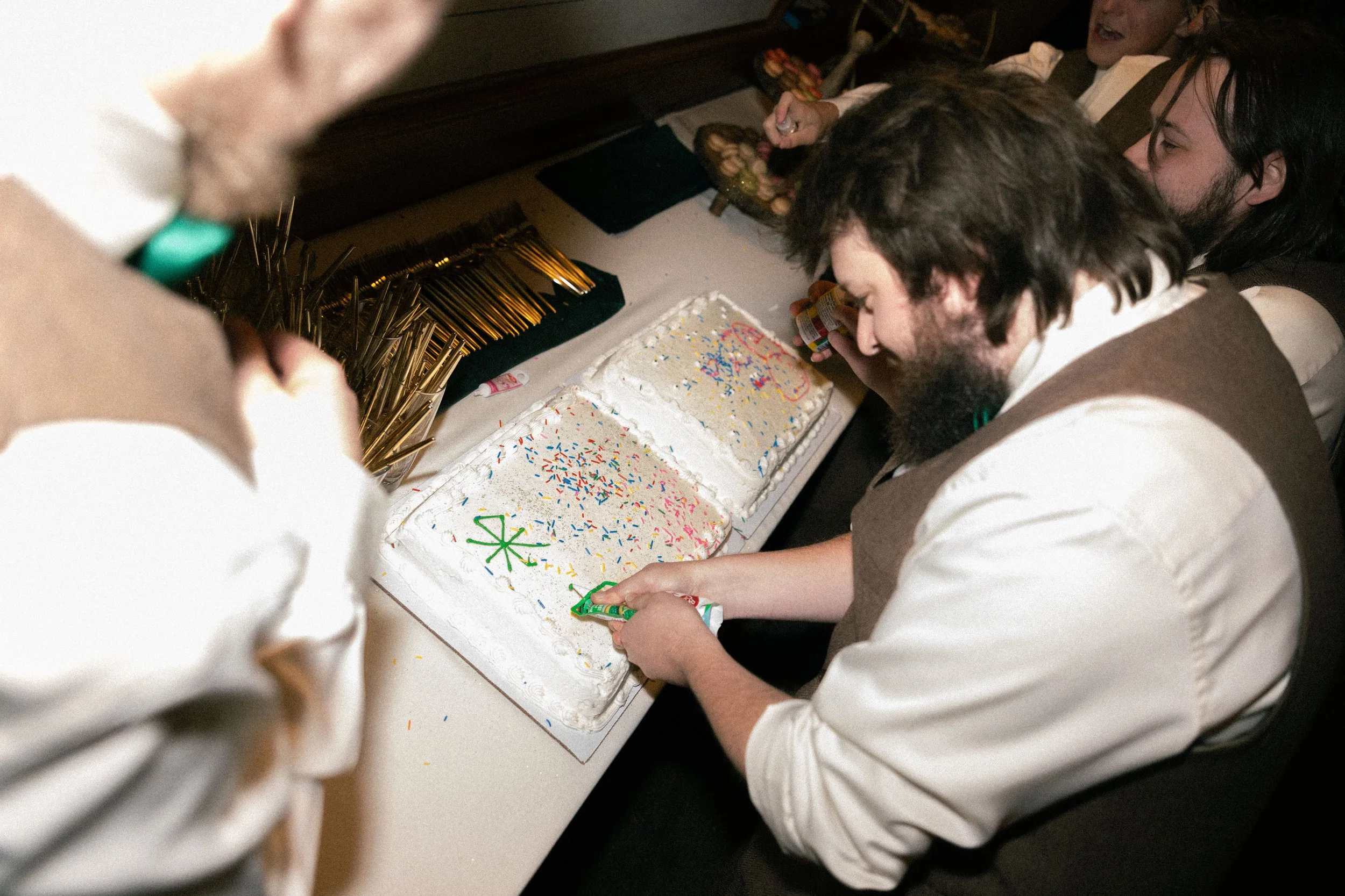 A person is cutting a birthday cake decorated with colorful sprinkles and icing, surrounded by others celebrating.