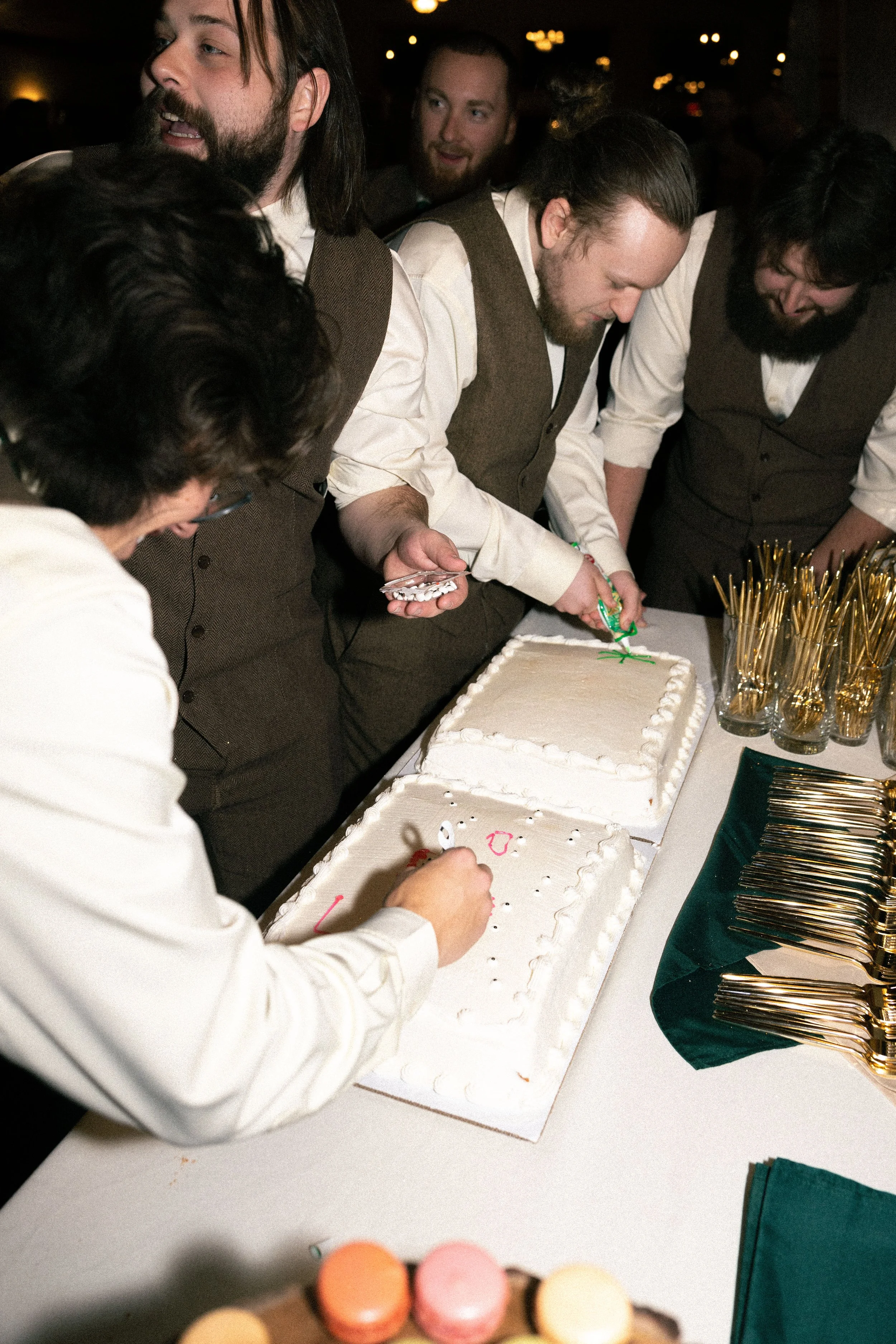Group of men in formal attire, gathered around a table with cakes, celebrating a special occasion with one man cutting or decorating a cake.