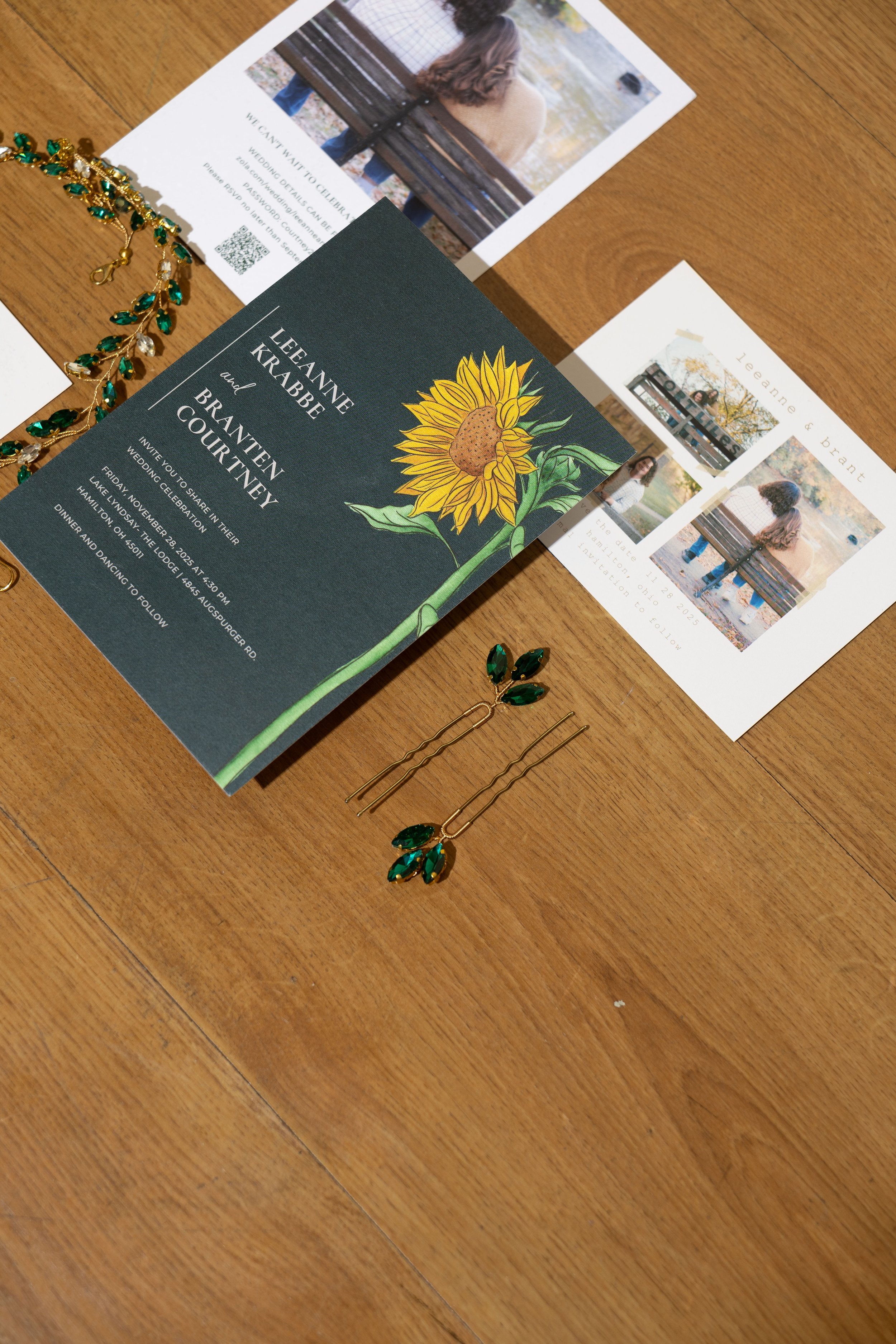 Wedding invitations and photographs on a wooden table, featuring a sunflower illustration and jewelry with green gemstone accents.