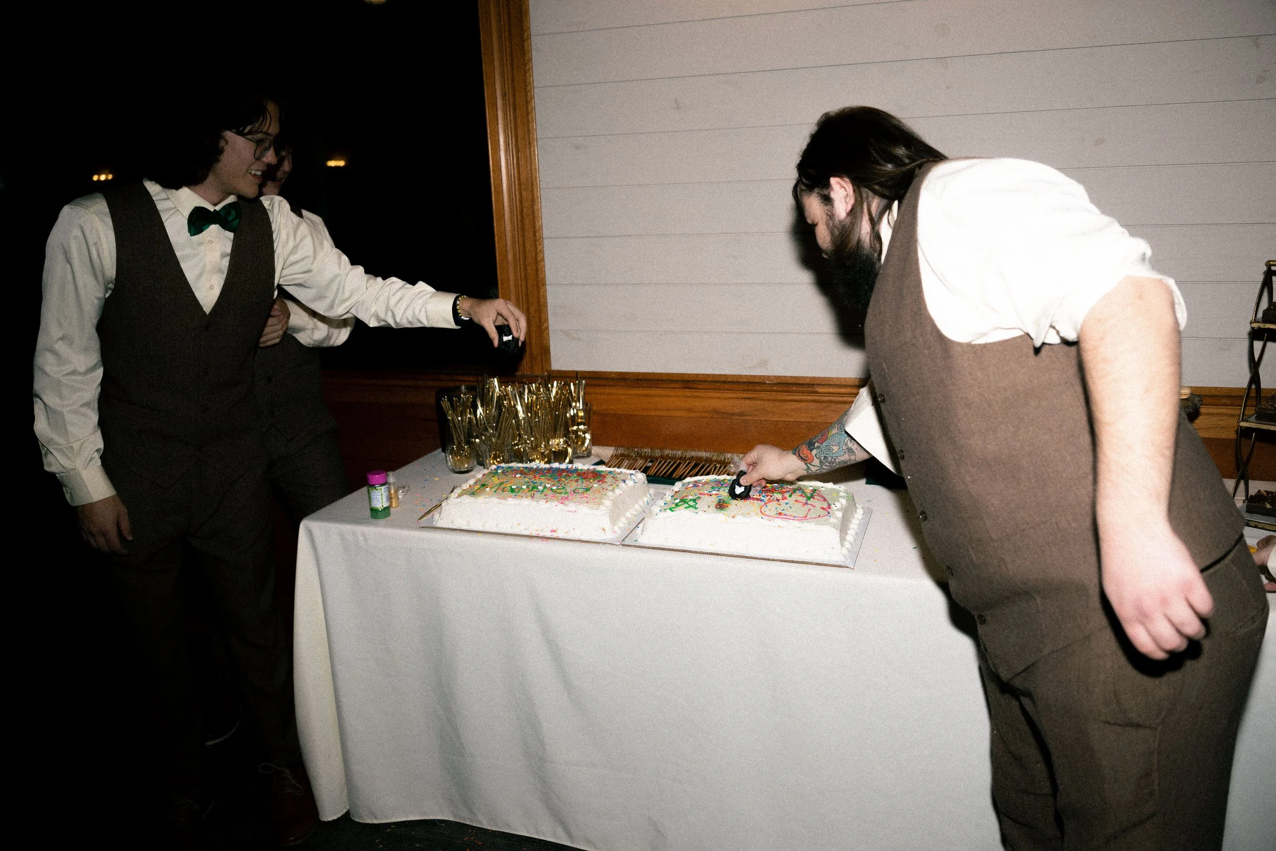 Two men, dressed in vests and shirts, are cutting two large birthday cakes with colorful sprinkles on a table. There are glasses and small bottles on the table, and a dark background.