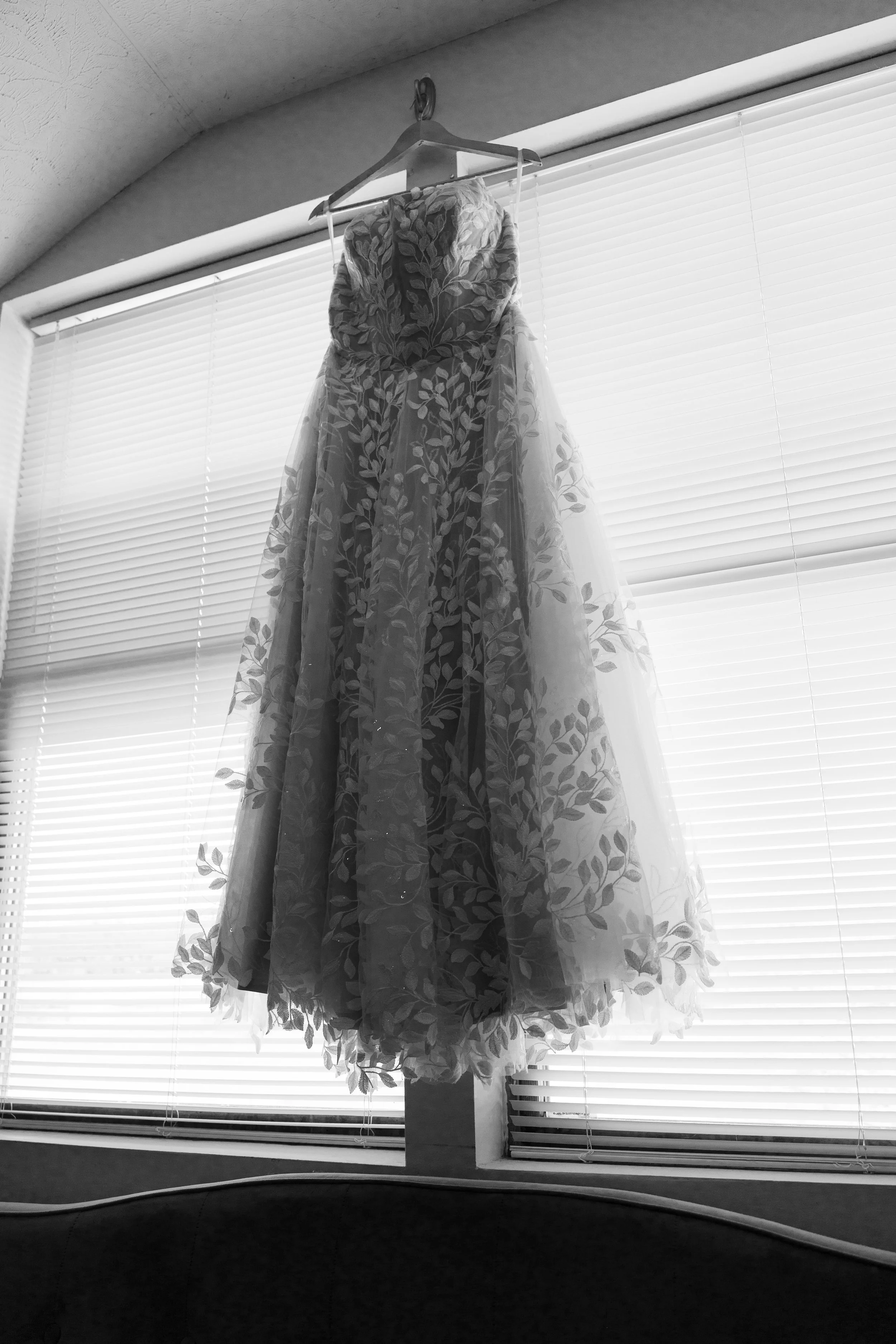 A floral wedding dress hanging on a hanger in front of window blinds.