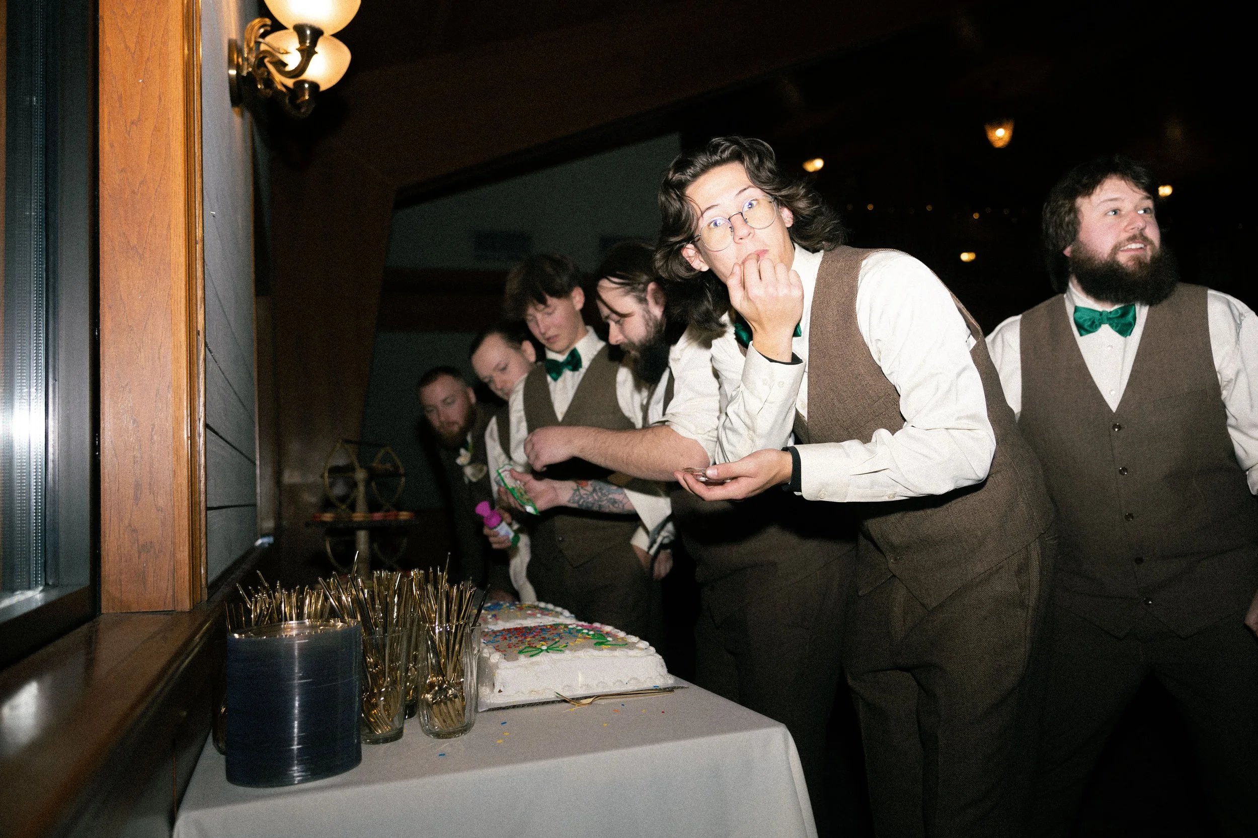 A group of six people dressed in vintage-style attire with vests and shirts, standing in line at a table with a decorated cake, gold utensils, and party supplies, at a party or celebration event.