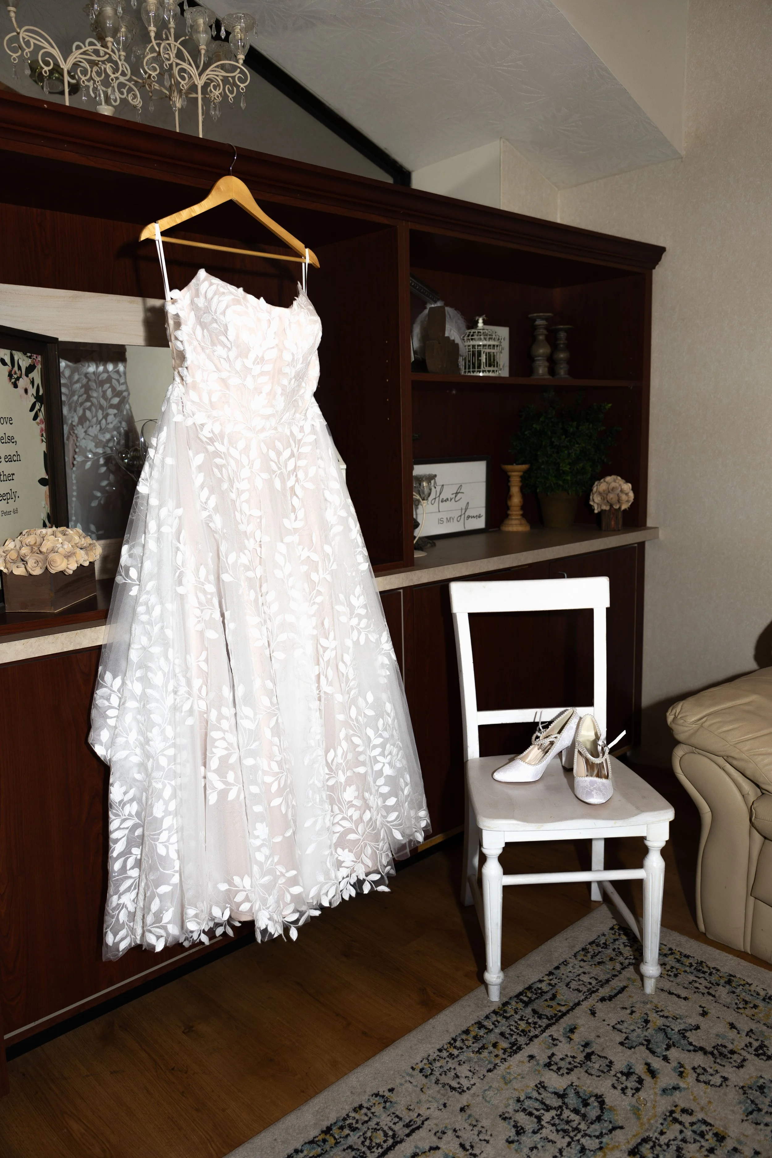 A white wedding dress with floral embroidery hanging on a wooden hanger, placed on a cabinet, white bridal shoes on a white chair nearby, in a living room with decorative shelves, a chandelier, and a patterned rug.