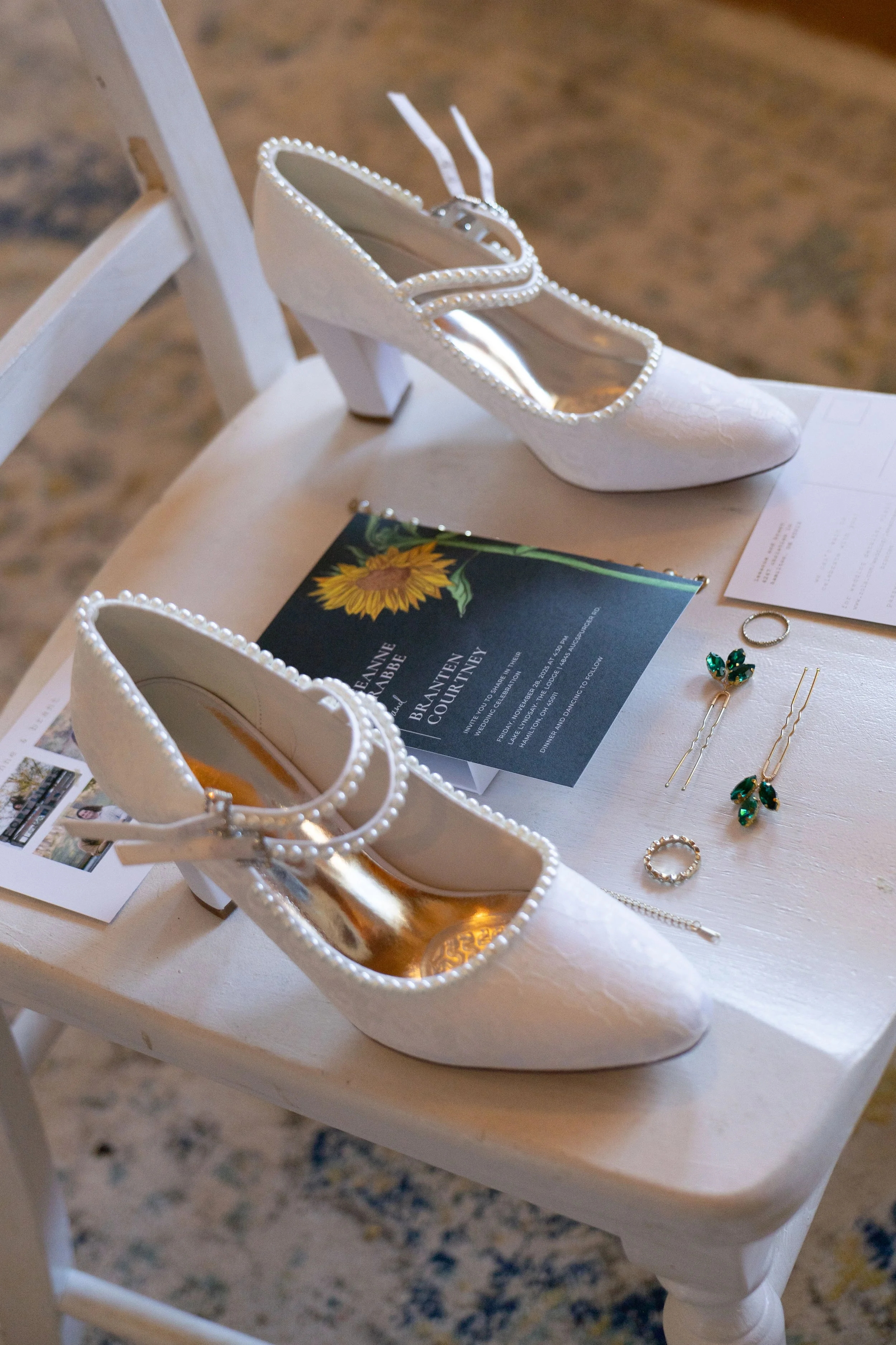 A pair of white bridal shoes with pearl embellishments on the heels and straps, along with a pair of green gemstone earrings, a ring, and a brochure on a white wooden chair.