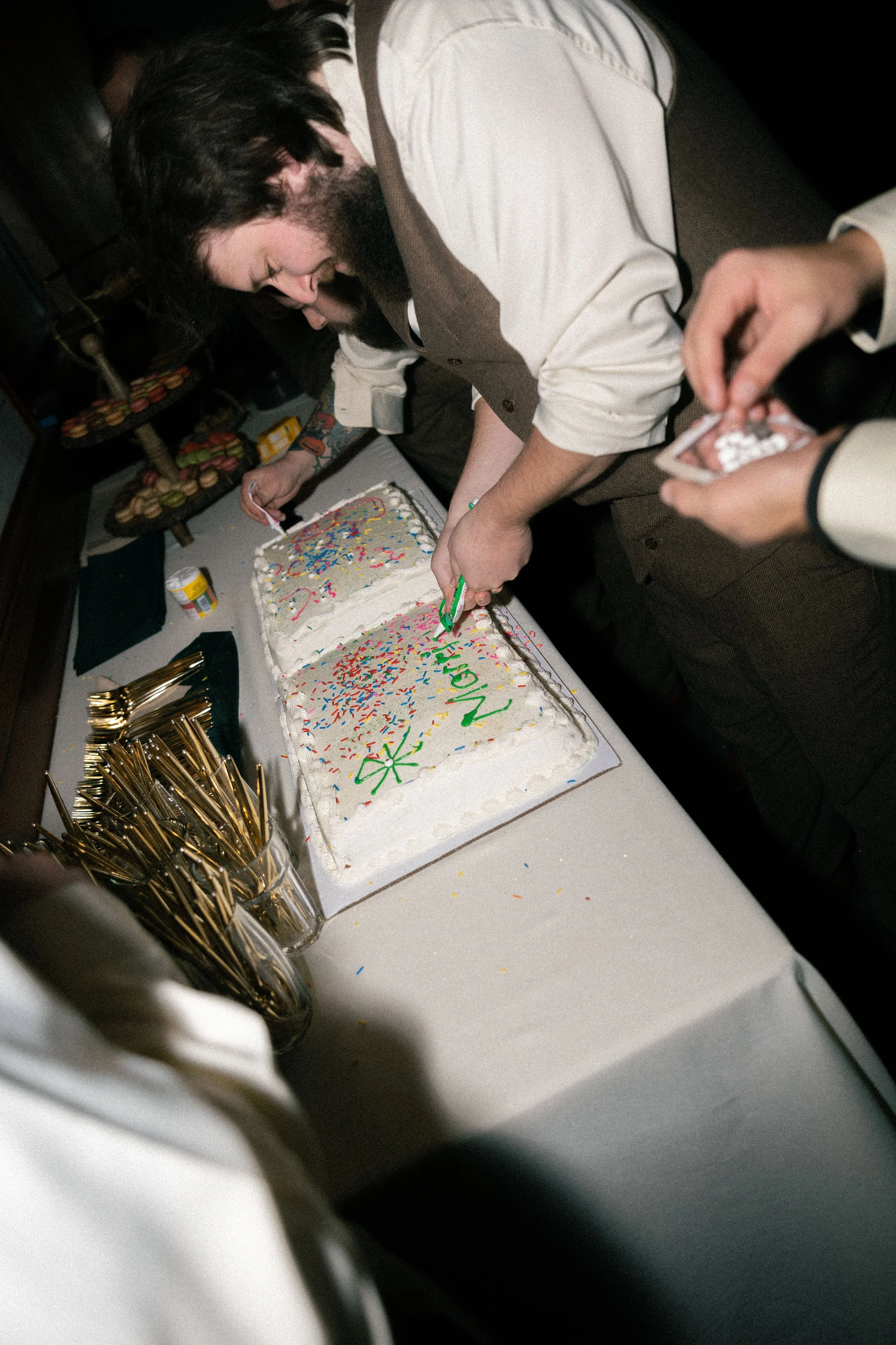 People cutting birthday cake decorated with colorful sprinkles with a frosting message that reads 'NICK.'
