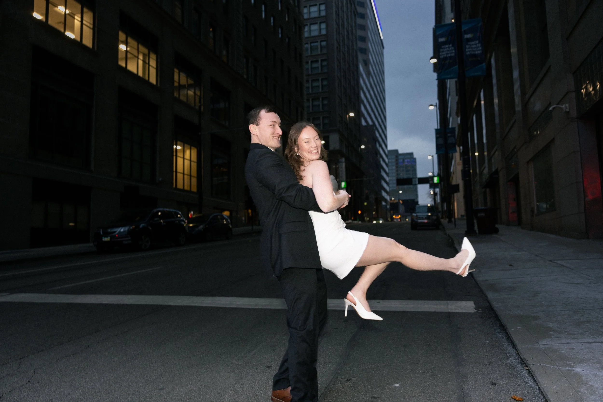 A man in a black suit lifts a woman in a white dress and high heels in an urban street at dusk, both smiling and enjoying the moment.