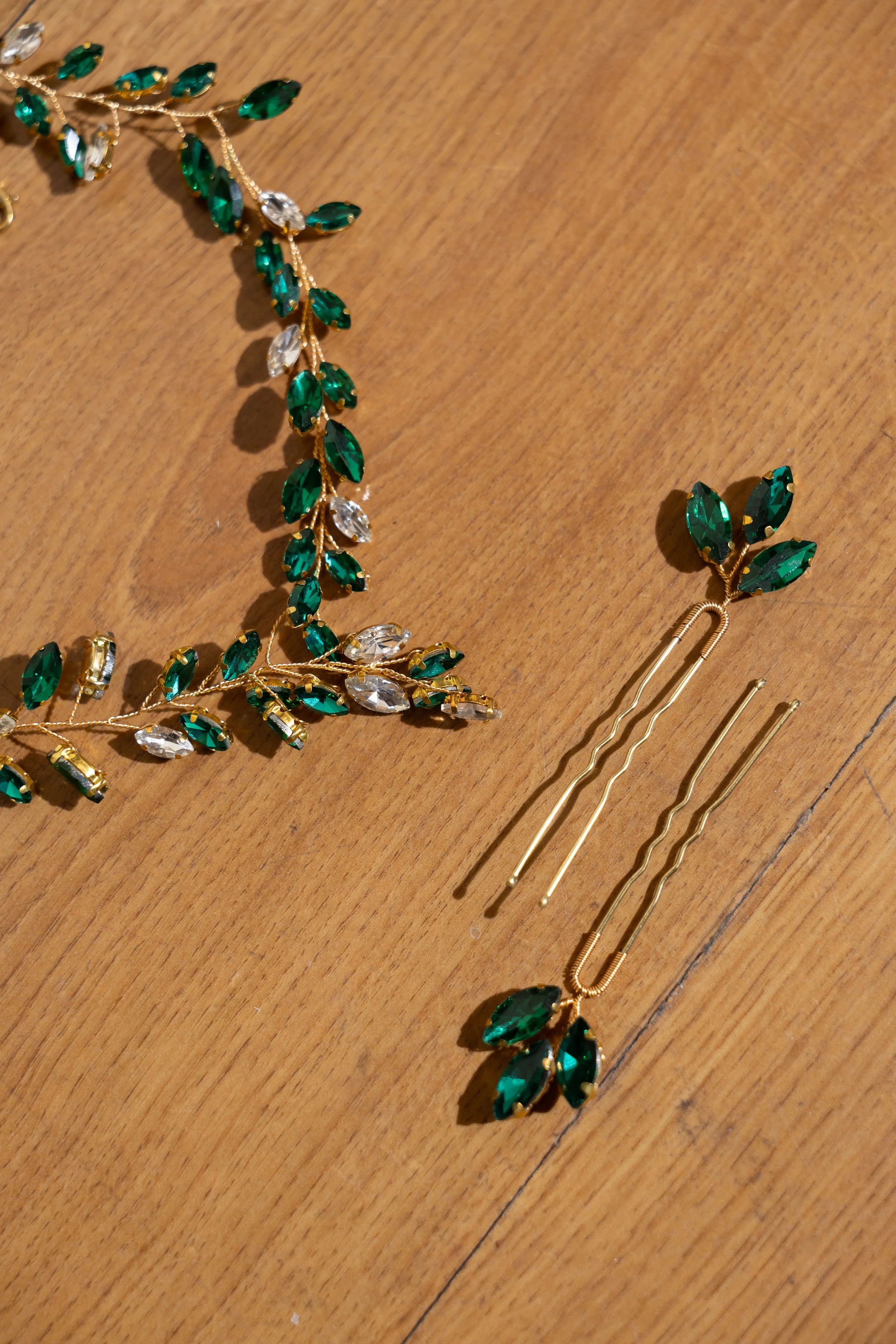 A set of emerald green and clear gemstone jewelry, including a decorative headband and hairpins, with gold-tone metal accents, laid out on a wooden surface.