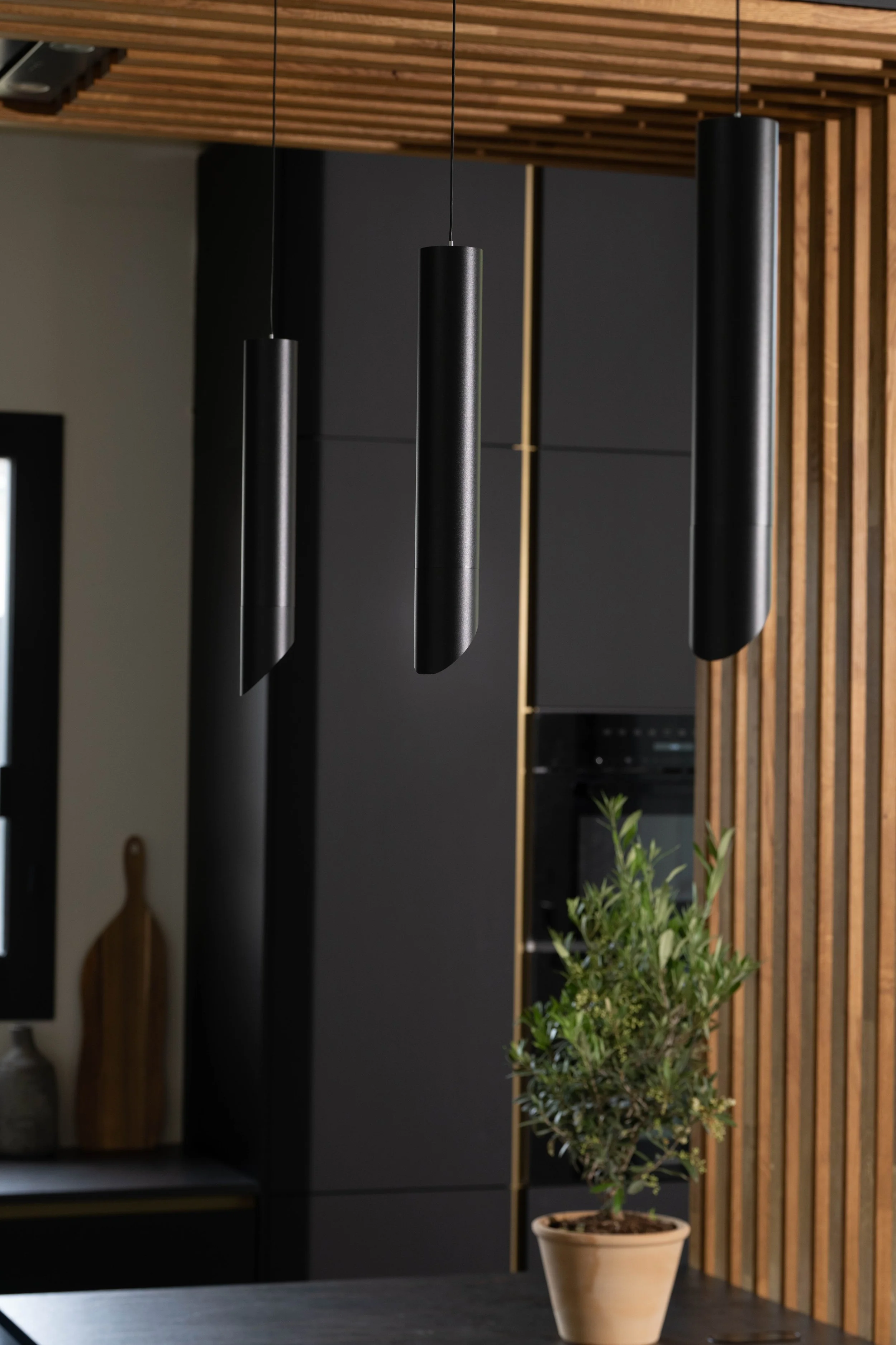 Modernt kök med tre svarta hängande lampor, ett träramvägg på höger sida och ett grönt växt i kruka på ett köksbord.