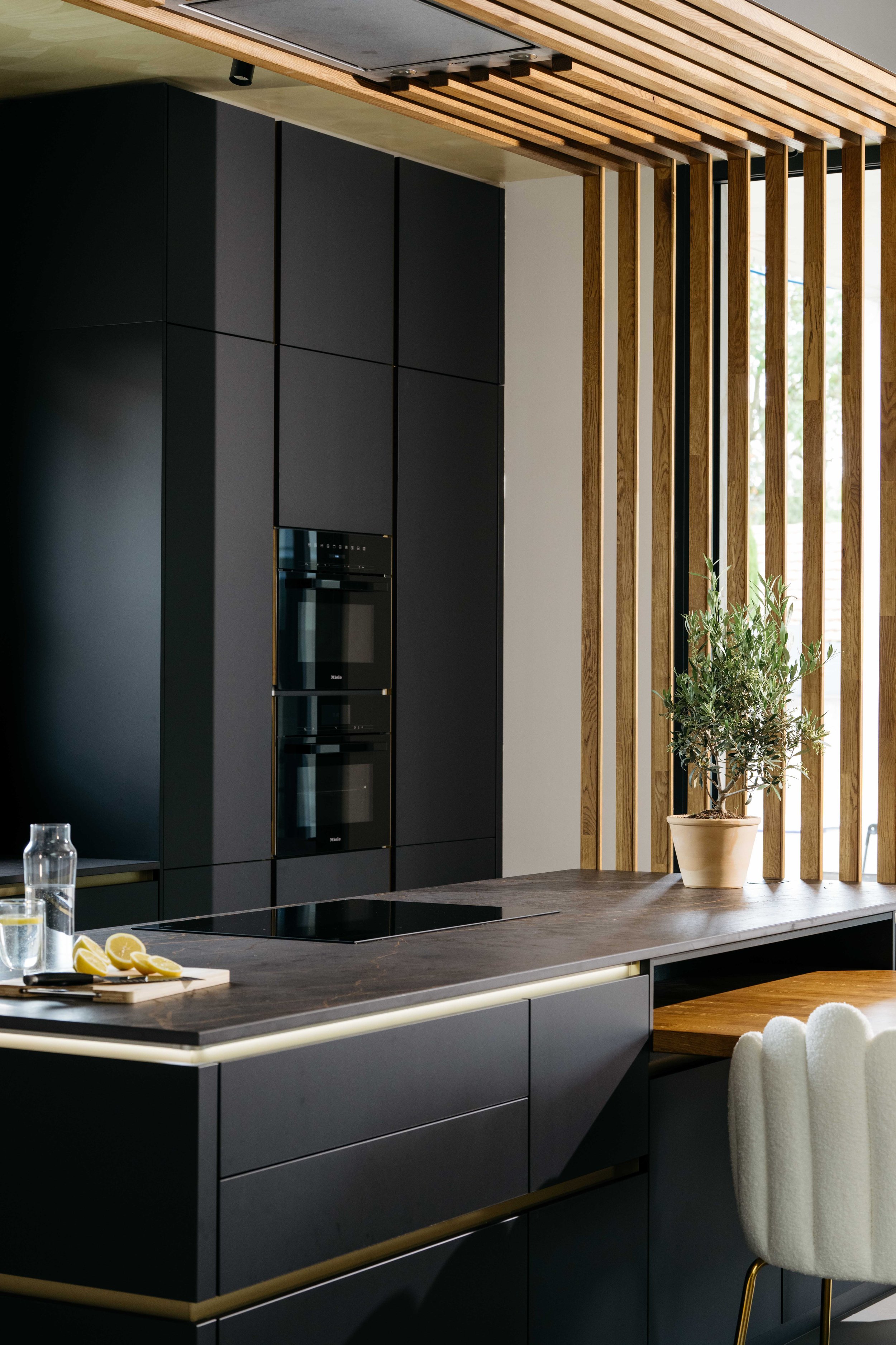 Modern köksinredning med svarta skåp, trädetaljer, en köksbänk med mörkt trä, en växt i kruka, ett fönster med vertikala trälister och några citroner på en skärbräda.
