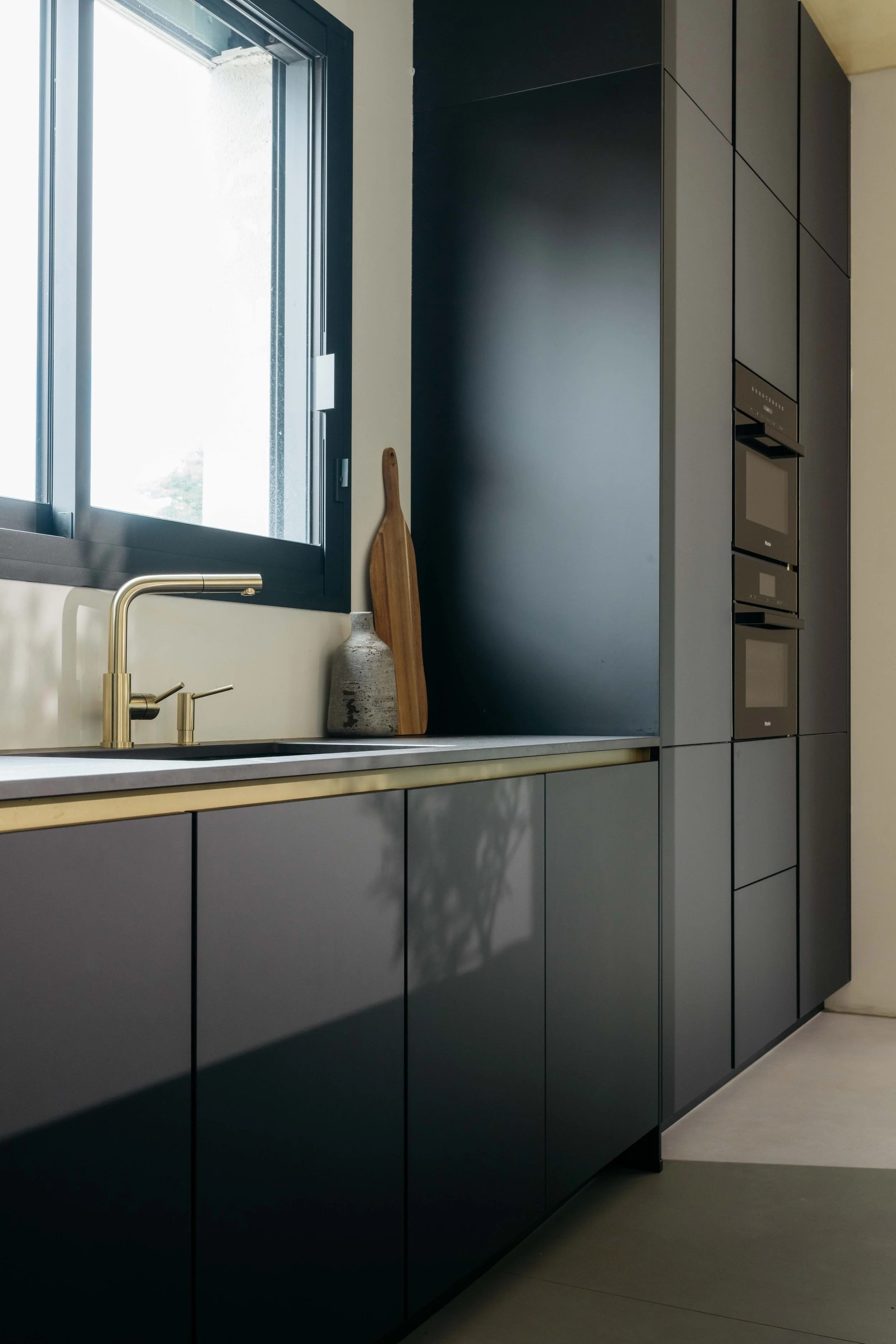 En modern kökssnickeri i mörkgrått med ett fönster, en diskho och ett bräde med dekorativa föremål, inklusive en keramikvas och en träskärbräda.