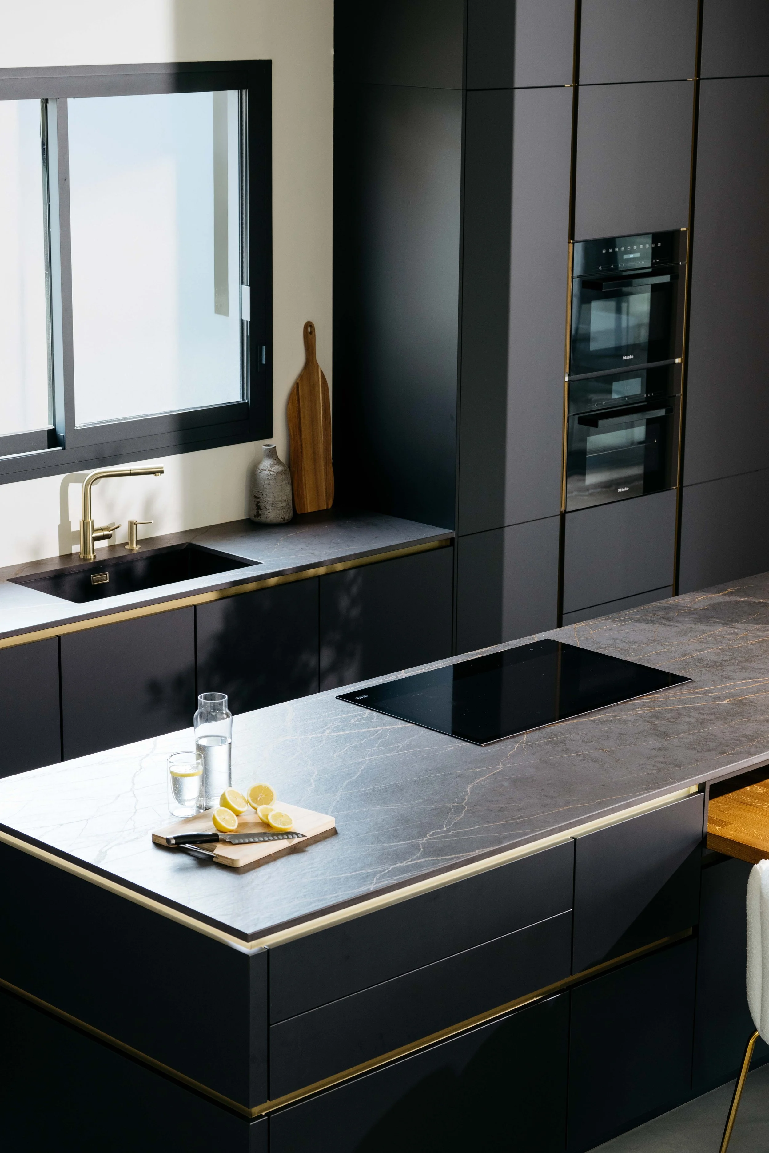 Modern köksinredning med svarta skåp, grå marmorbänkskiva, svart kran och ugn, köksbord med citroner och vatten.