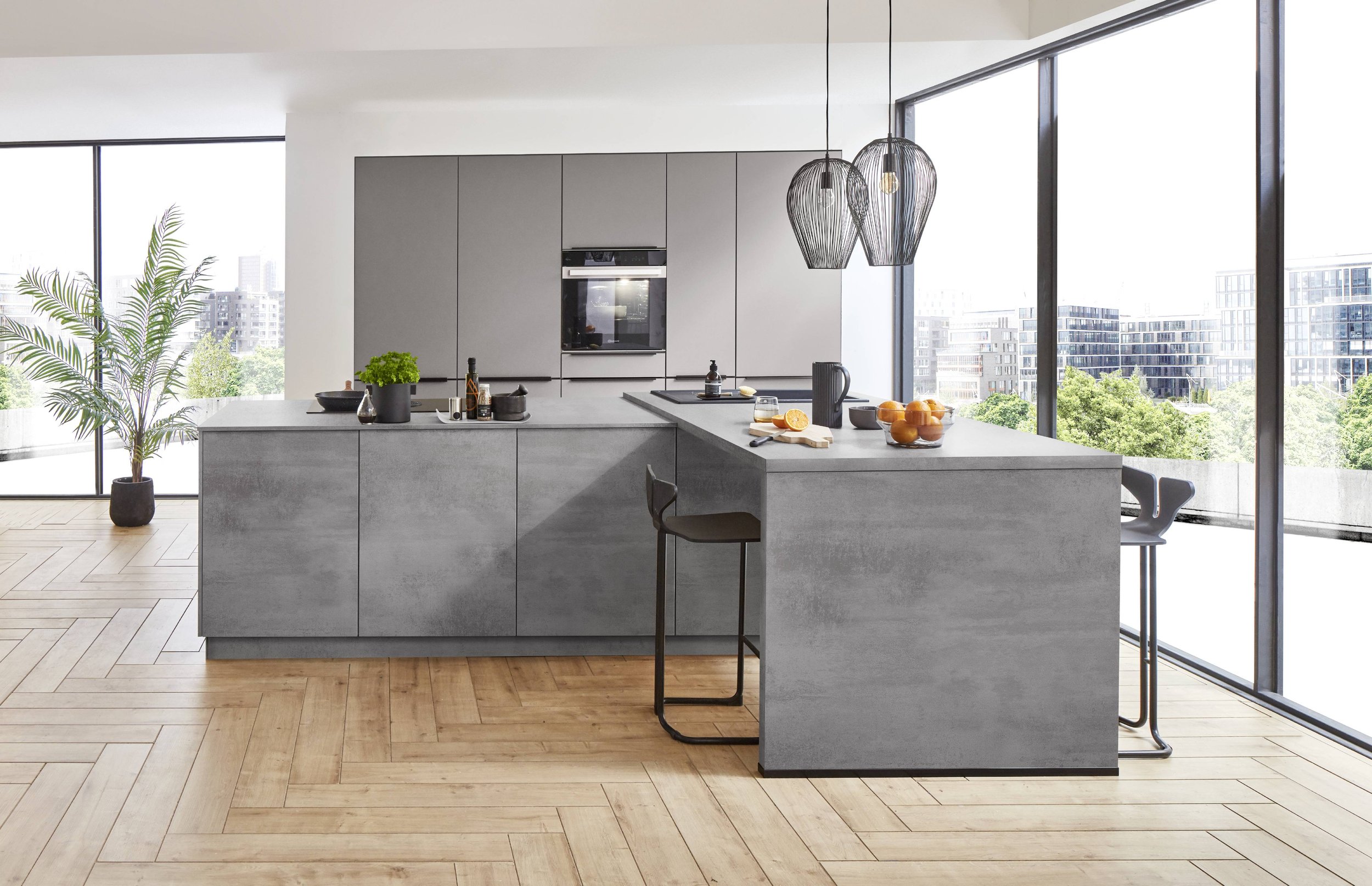 Modern köksö i grå betong med barstolar, stora fönster med utsikt över stadens byggnader, träd och tak, samt köksutrustning och frukt på bänken