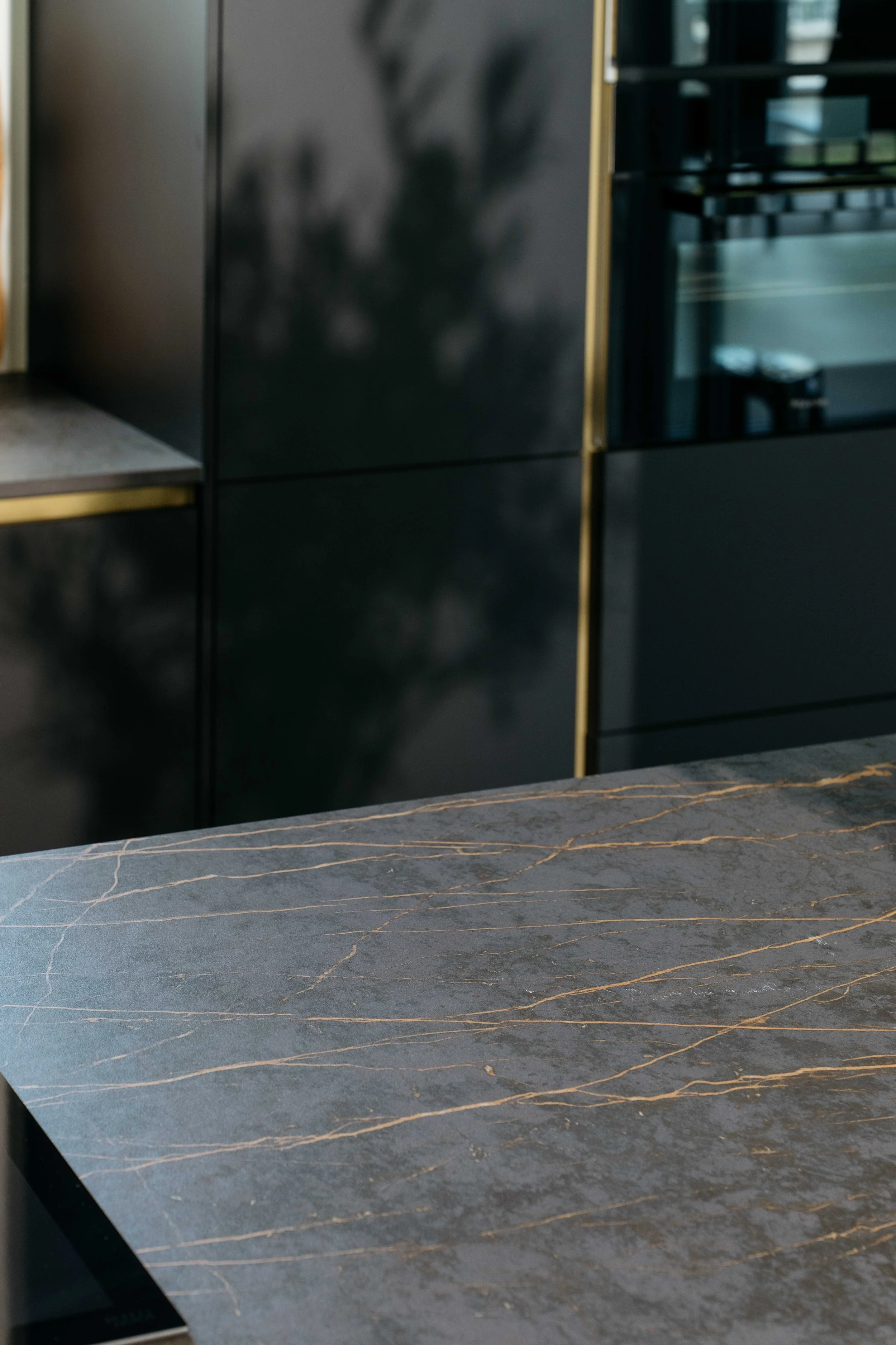 En köksbänk i mörk sten med guldfärgade ådringar, i bakgrunden svarta skåp och ett glasdörrskåp.