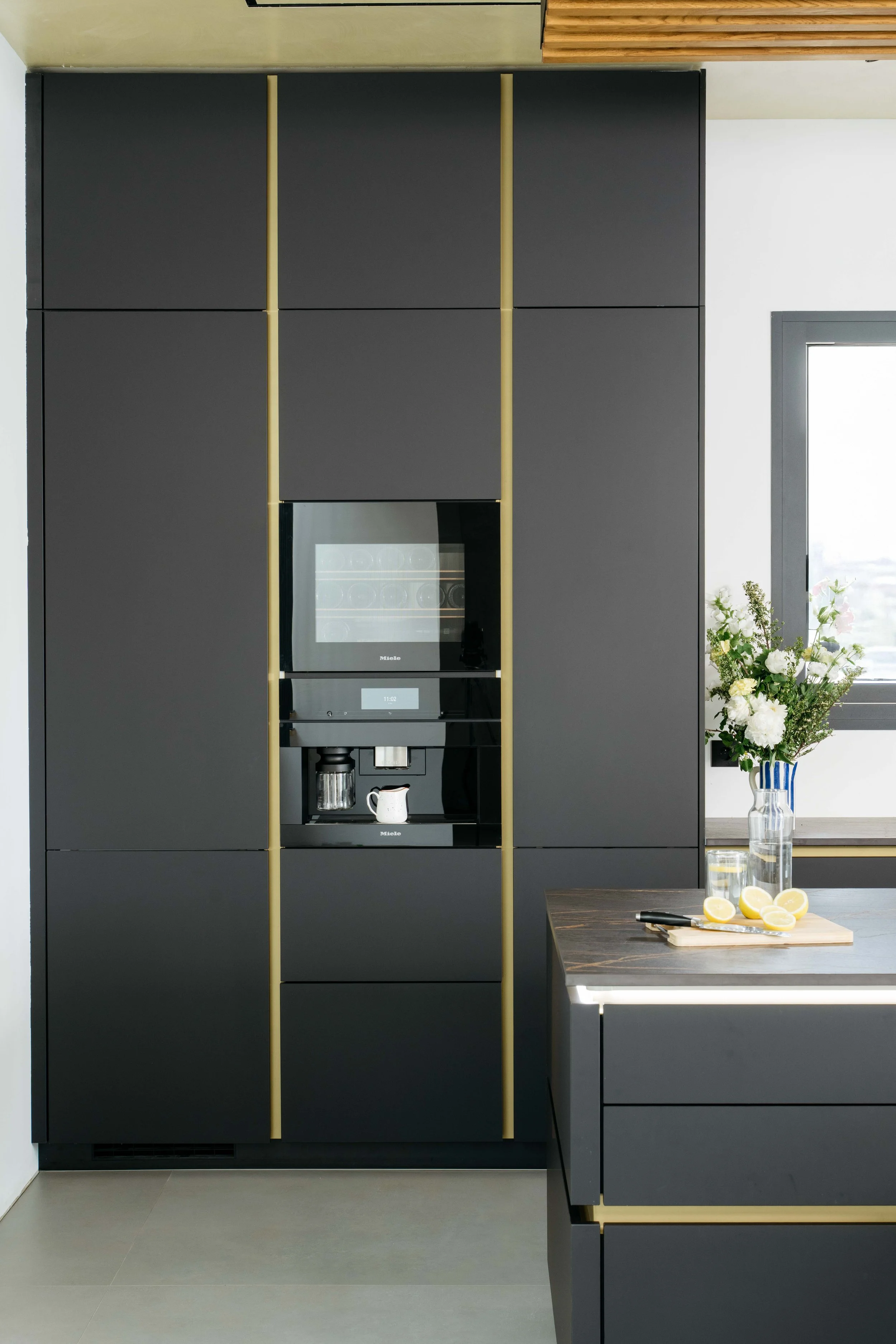 Svart köksskap, integrerad kaffemaskin och mikrovågsugn, samt köksbord med citroner och en blomsterarrangemang.