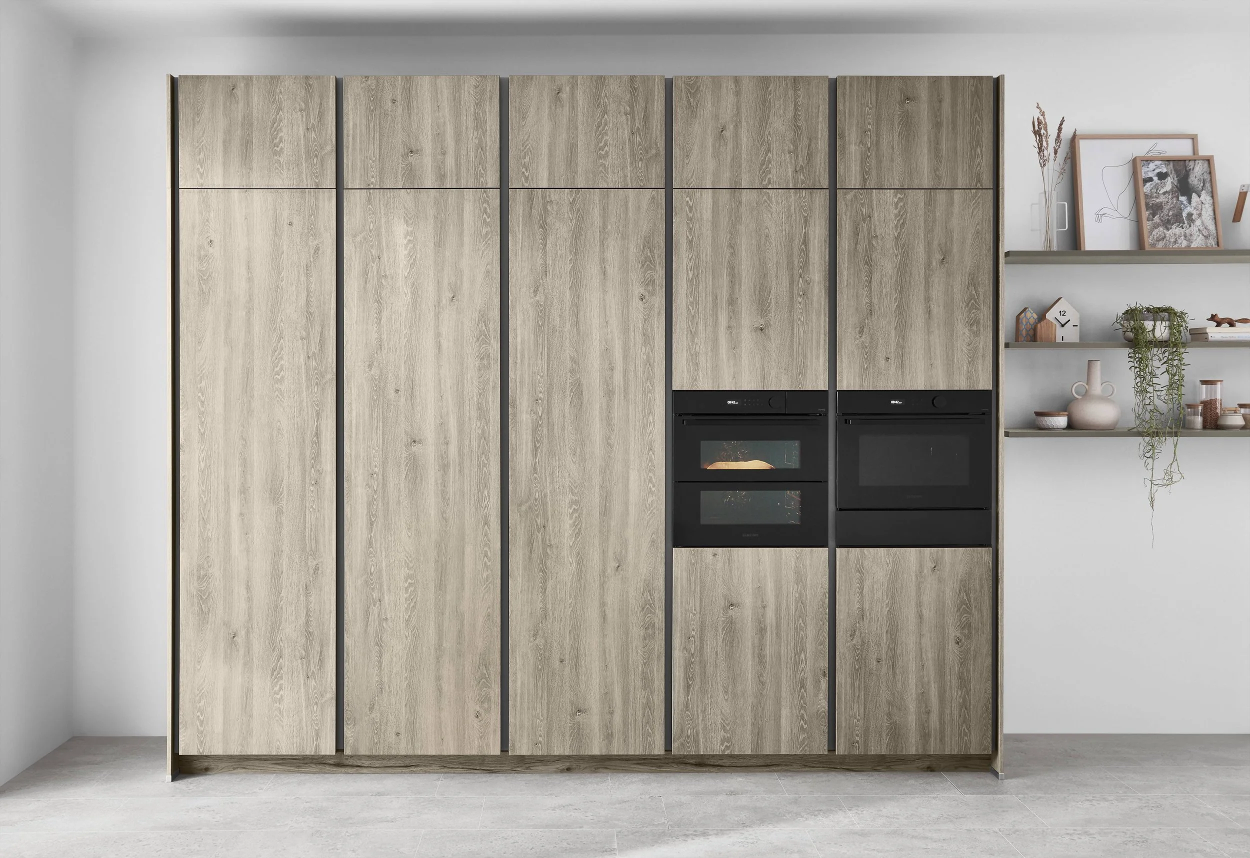 Kök med inbyggda svarta ugnar och skåp i ljus träfinish, vita väggar och vägghyllor med dekorationer och gröna växter.