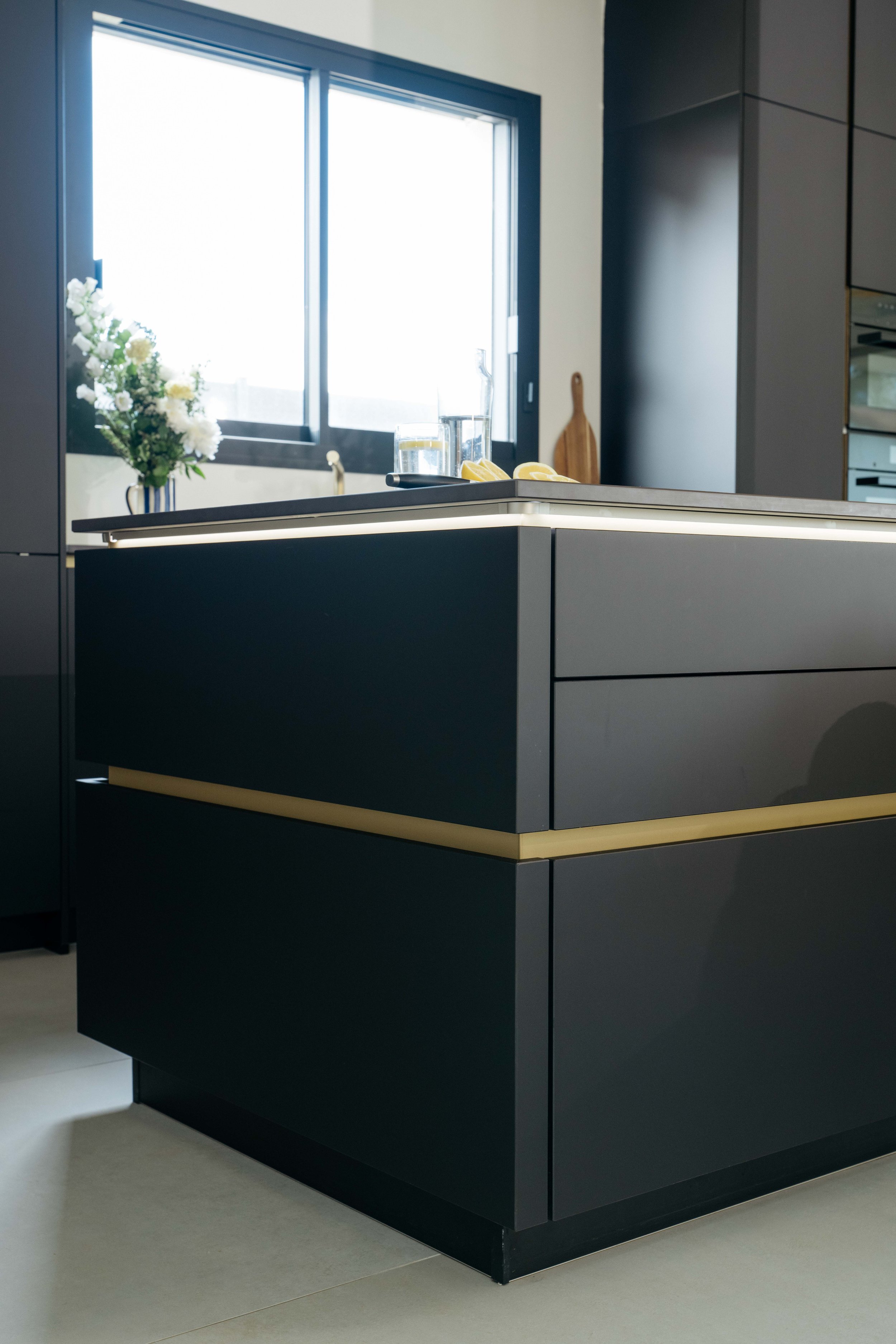 En modern köksö i svart med ljus listdetalj, mot ett fönster med blomsterarrangemang och köksredskap på bänkskivan.