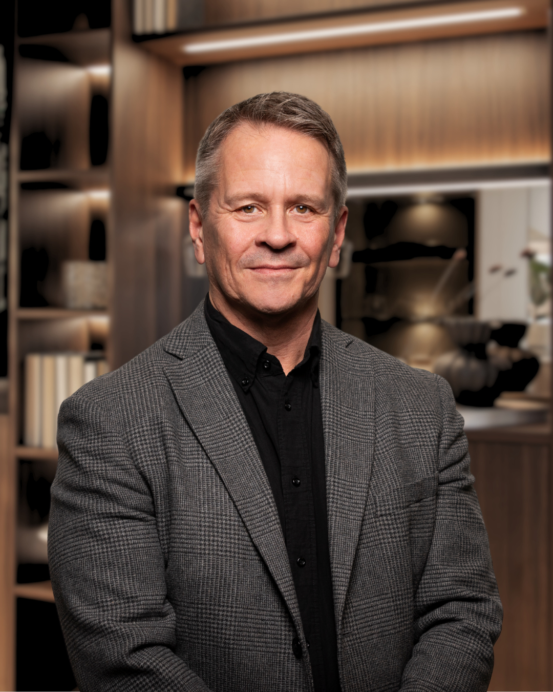 En man i kostym och mörk skjorta står framför en bokhylla med böcker och dekorativa föremål.