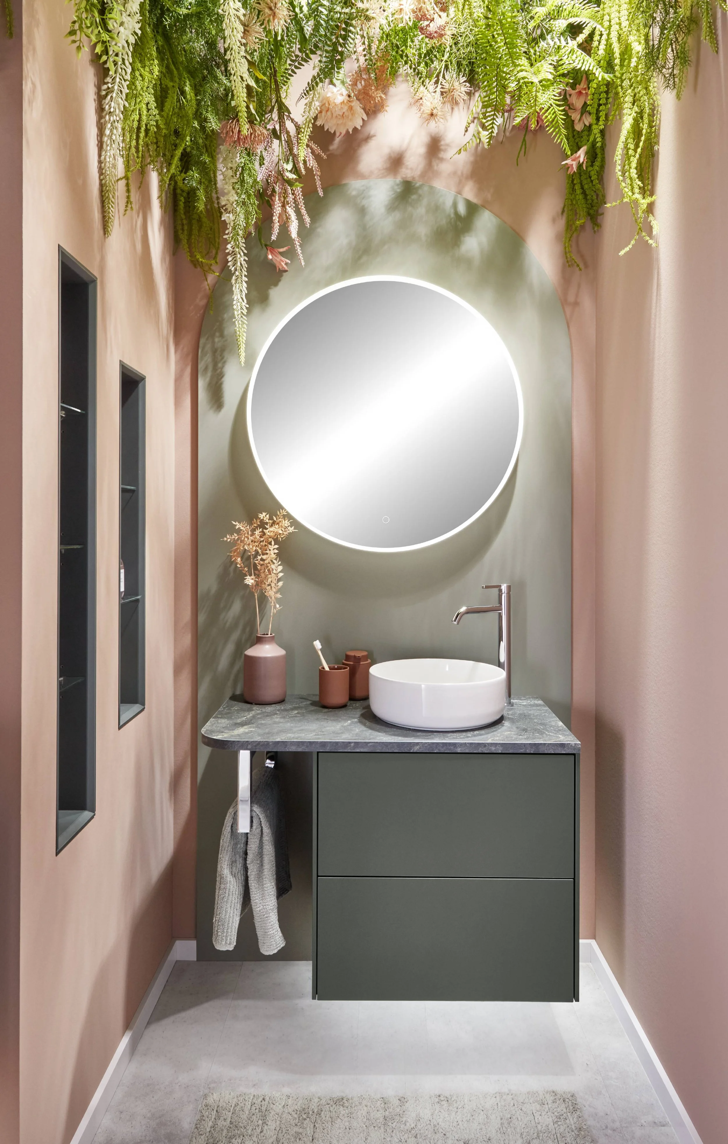 Snygg badrumsinredning med en rund spegel med belysning, vit handfat på en mörkgrön bänk, ett prydnadsvas med torra växter och växter ovanför spegeln, rosa väggar och vardera sidan hyllor med små föremål, samt en handduk hängande på sidan.