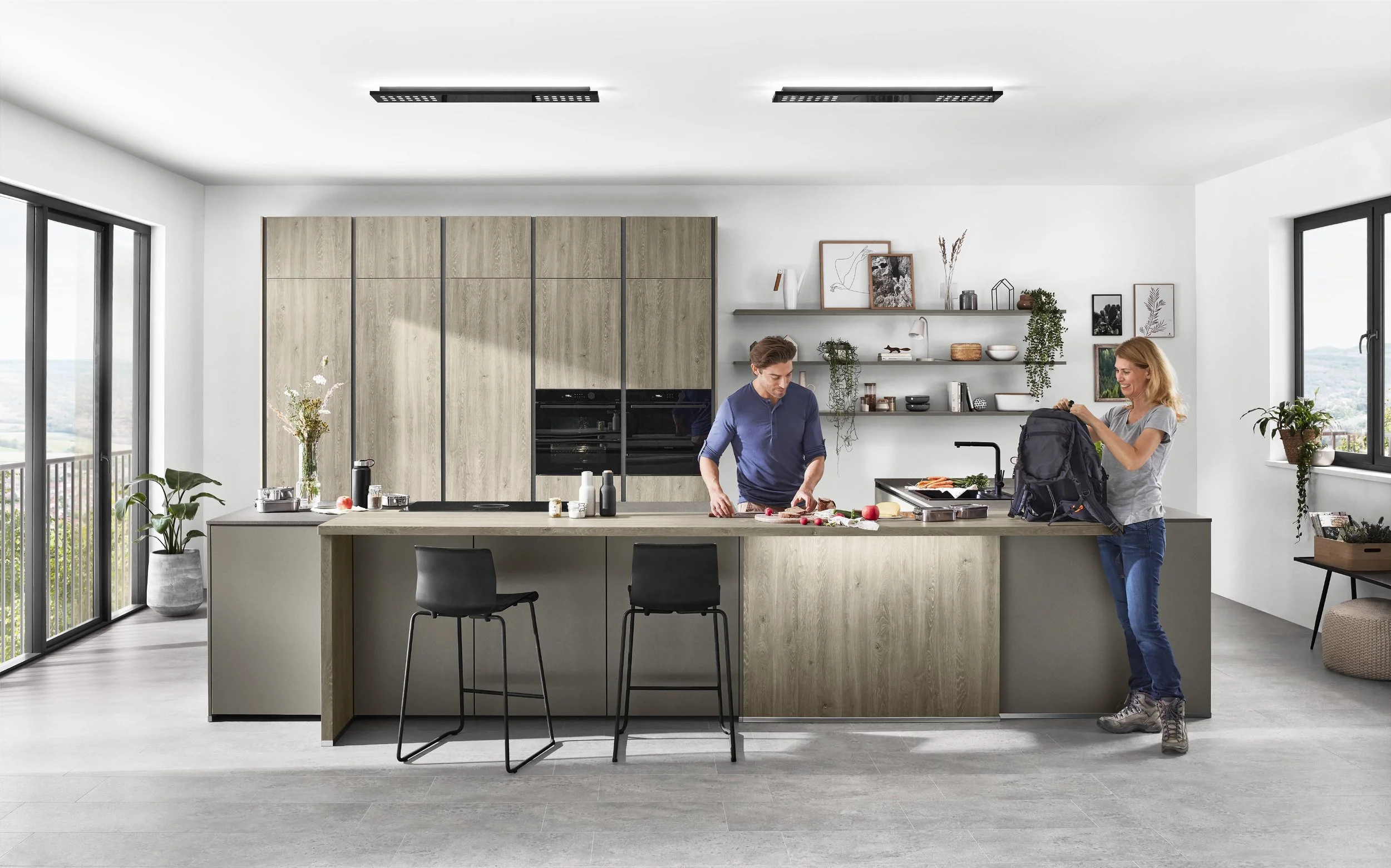 En modern köksscen med två personer, en man står vid köksbänken och skär grönsaker, en kvinna packar en ryggsäck, stora fönster, minimalistiska hyllor och växter.