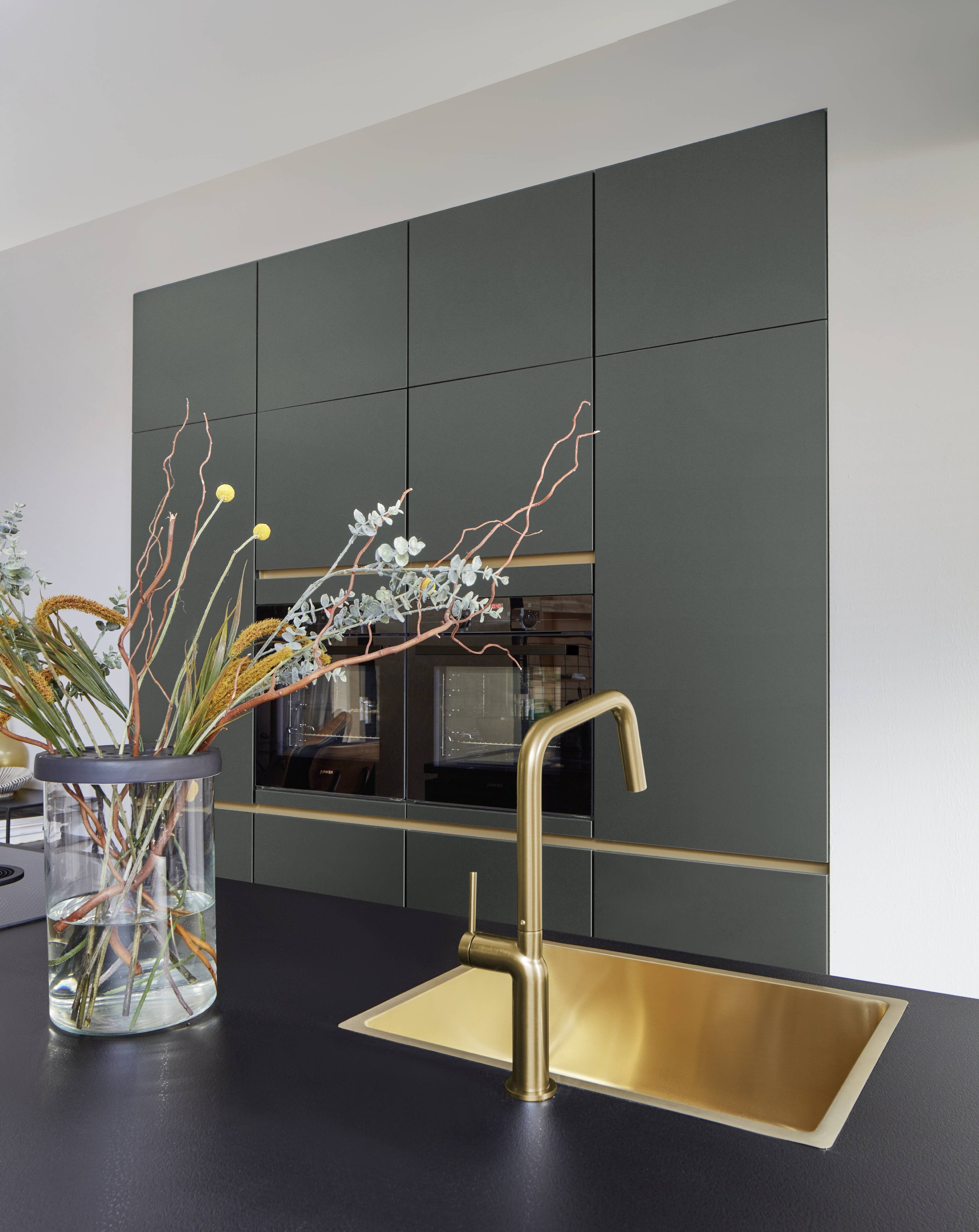 Moderne kök med gröna skåp, gulddetaljer, svart bänkskiva, guldfärgad diskho och blandare, samt en vas med blommor.