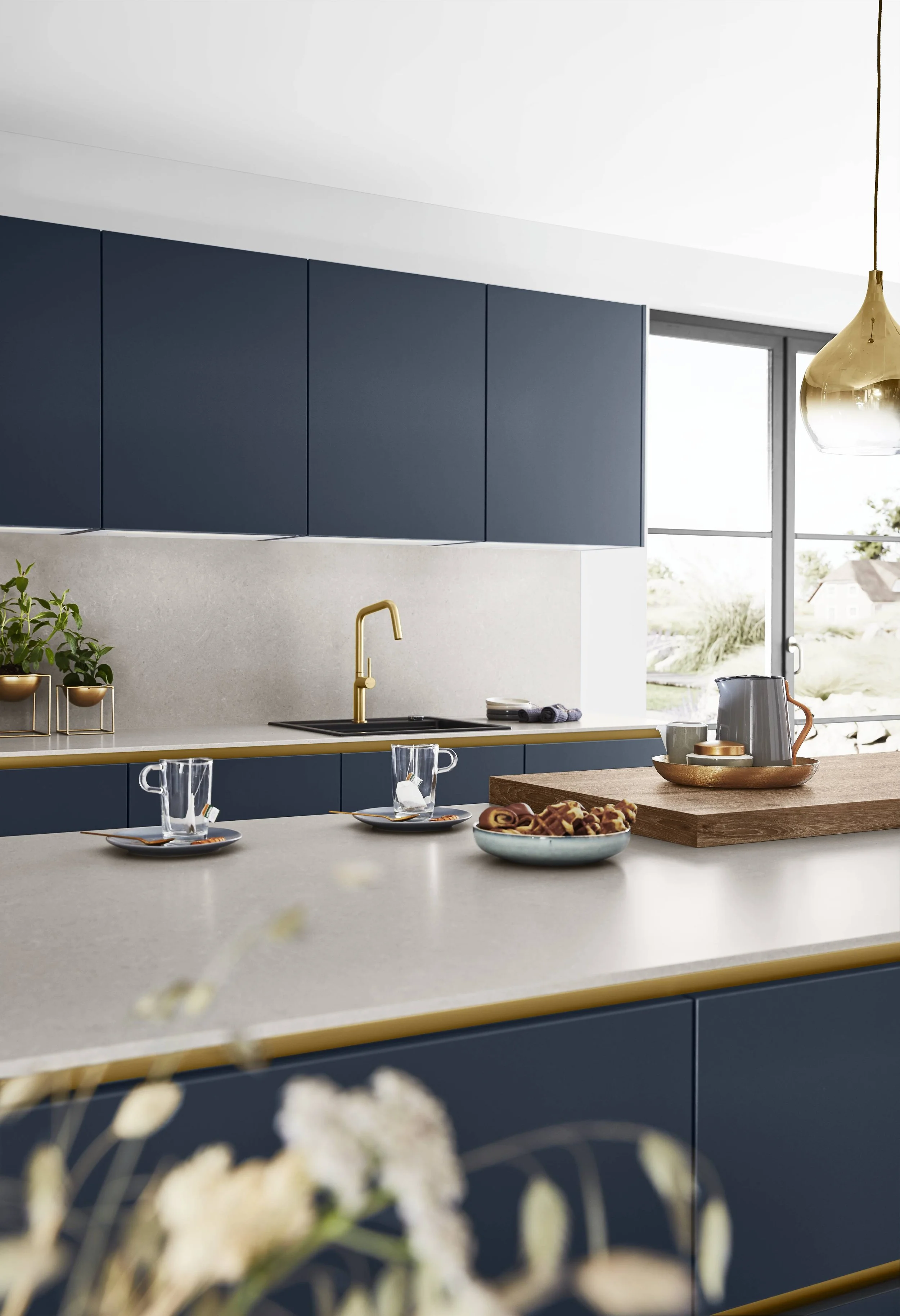 Modernt kök med blå skåpsluckor, beige bänkskiva och gulddetaljer, stor fönstervägg och ett blickfång i guld.