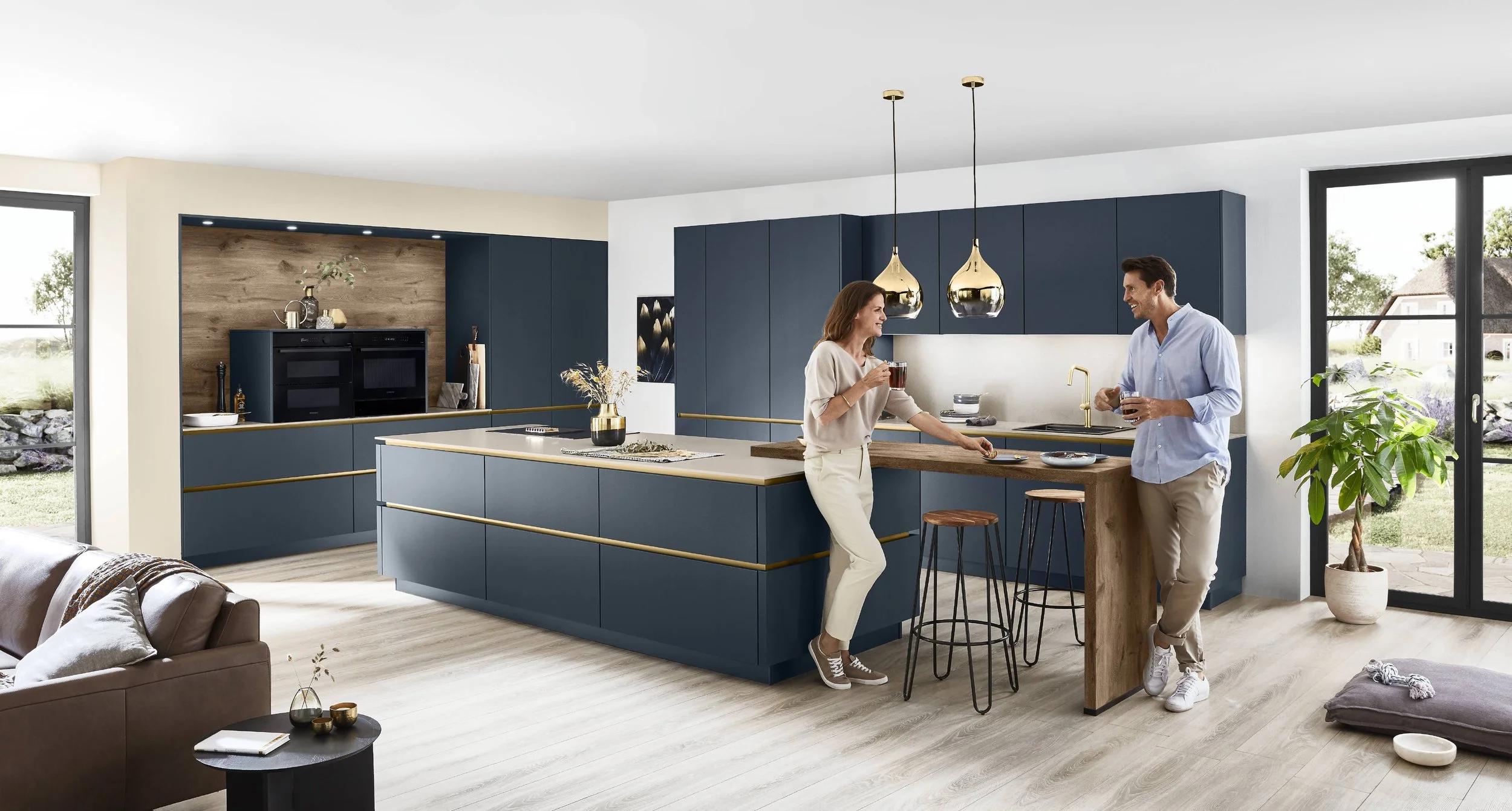 En modern köksscen med två personer som pratar och dricker te, blå skåp, hemmasnickrad köksö i trä, stora fönster, gröna växter.