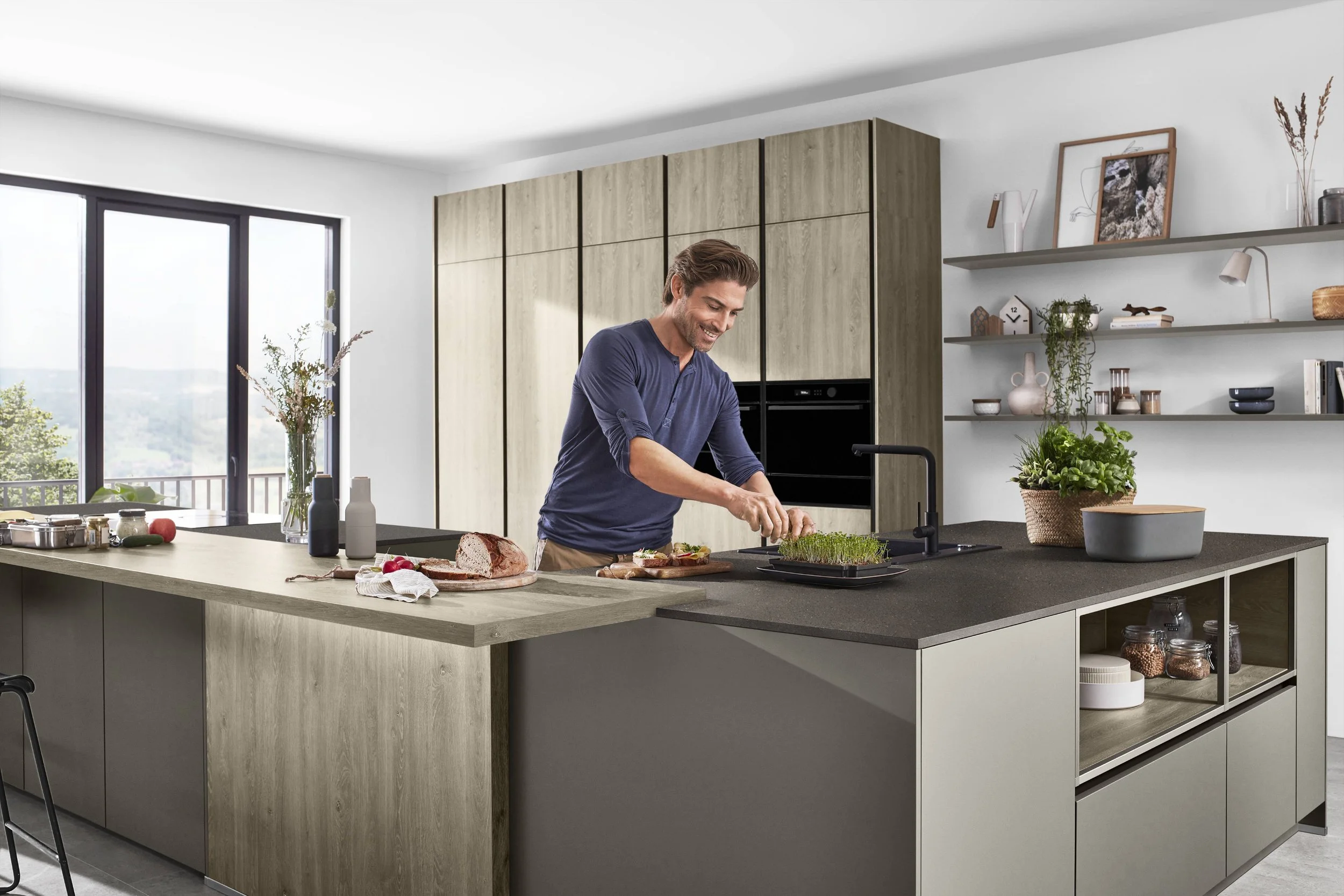 En man som lagar mat i ett modernt kök med stora fönster, grönska utanför, och hyllor med dekorationer och kokböcker.