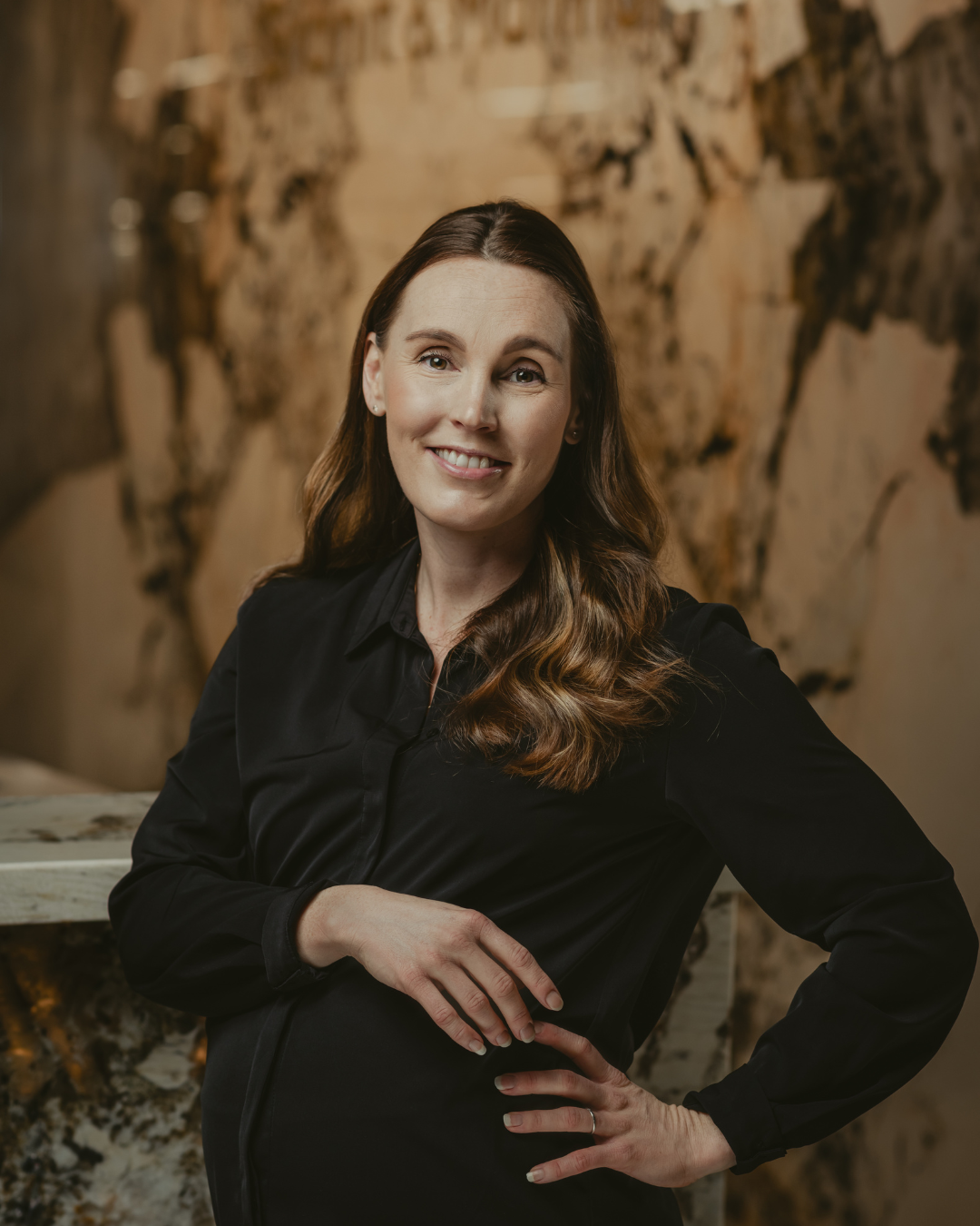 En kvinna med långa, bruna lockigt hår, klädd i en svart blus, står framför en konstnärlig vägg med ett världskarta. Hon ler mot kameran.