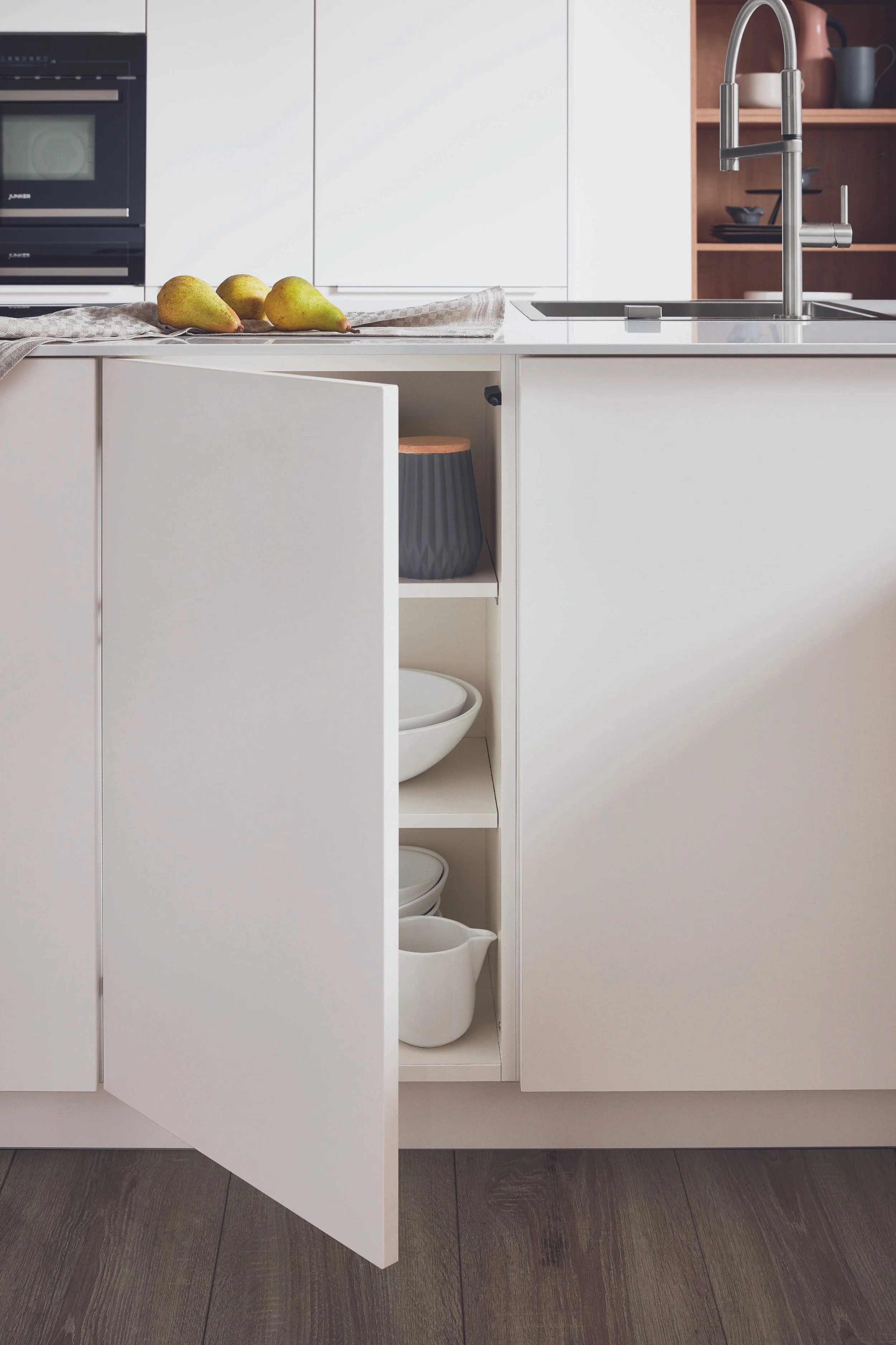 Vit köksö med öppen skåp som innehåller keramikskålar och kannor. På köksbänken finns tre päron och en trasa. Svart diskho och kökskran. Bakom syns vita skåp och ett glasparti.