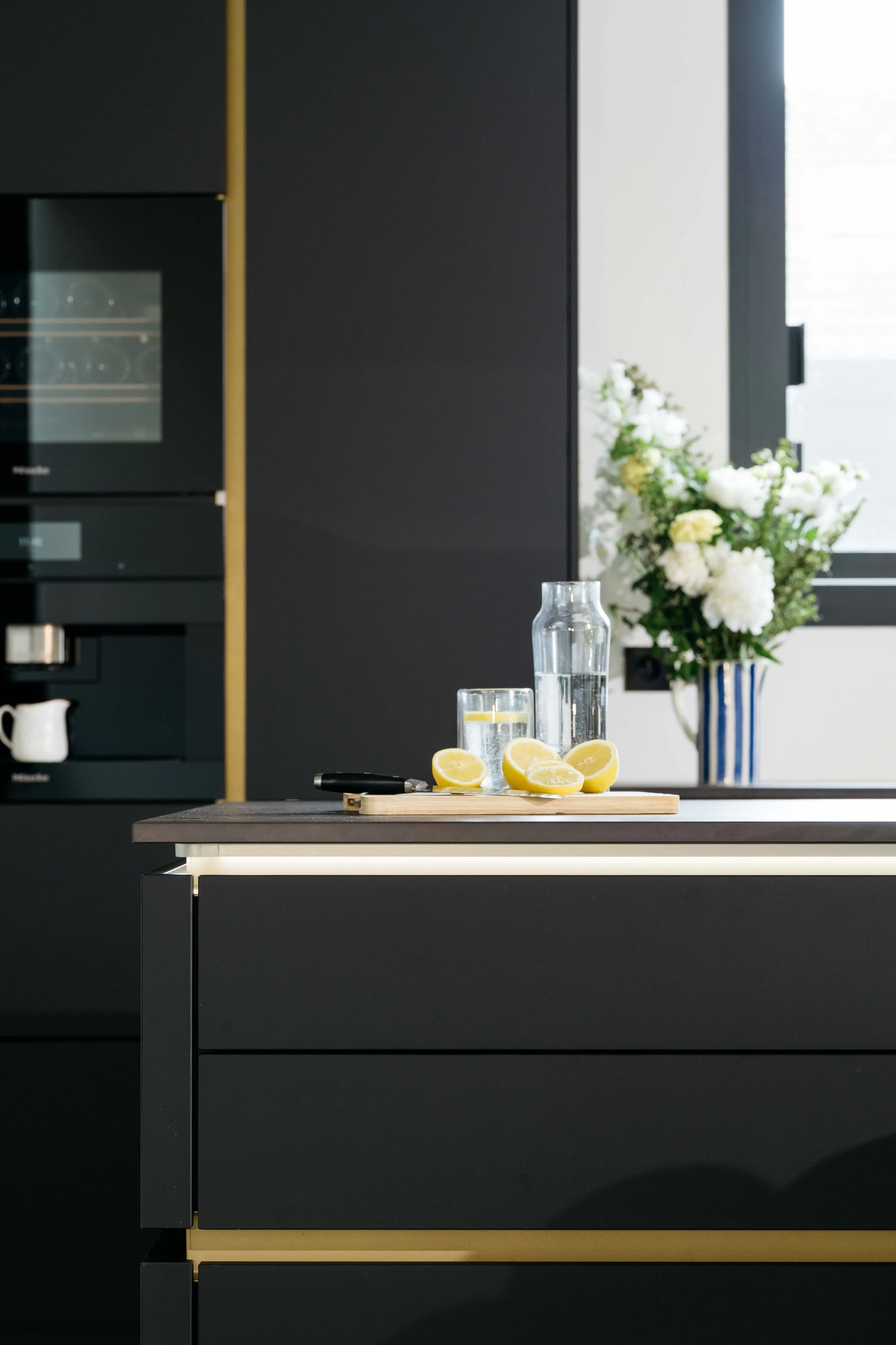 Kök med mörk skåp och ett köksbord med vatten, citronskivor och en kniv, samt en blå och vit blomkruka med vita blommor.