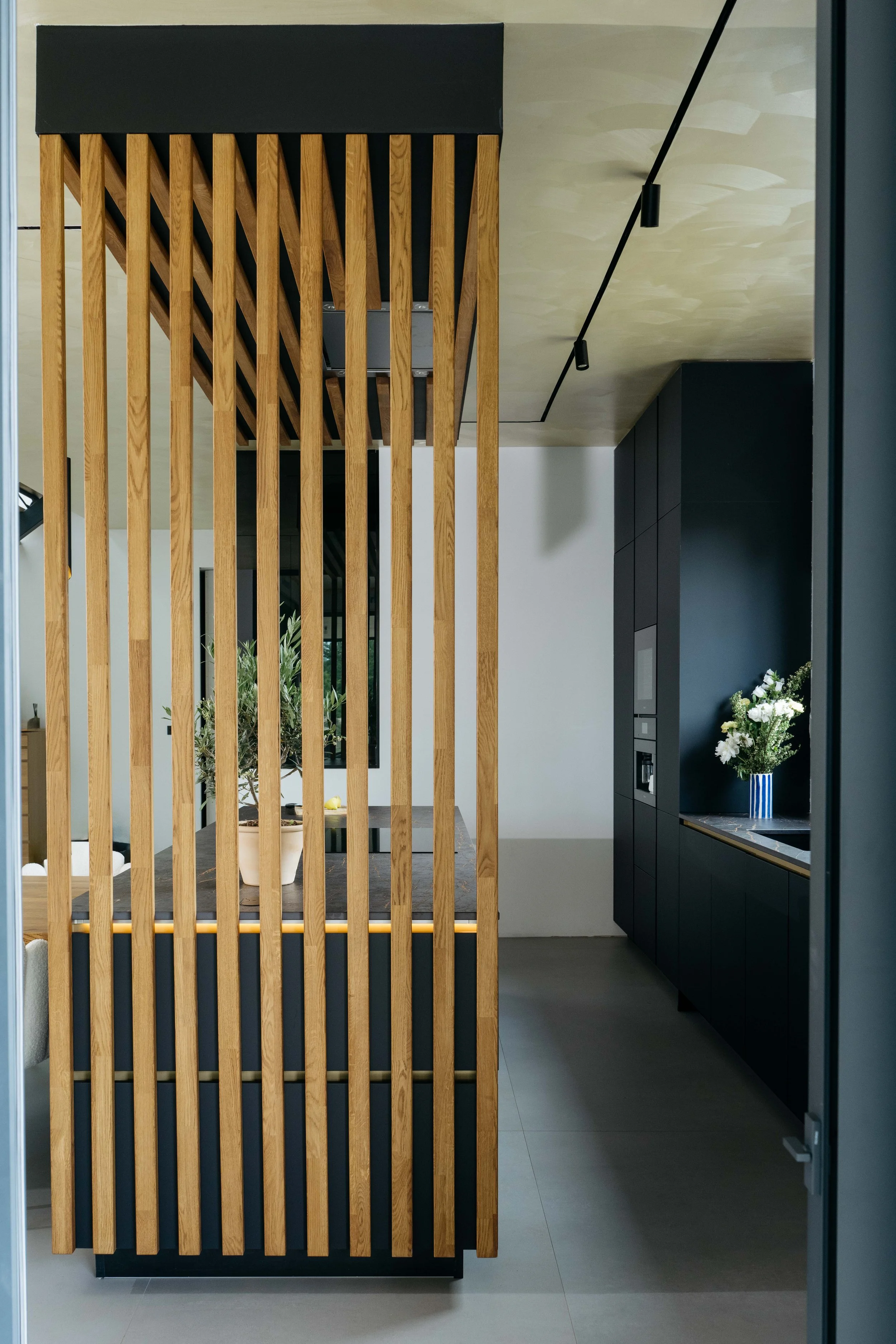 Interiör av ett modernt kök med svarta skåp och en träskärm som delar av köket. Det finns en blombukett i en blåvit vas på köksbänken.