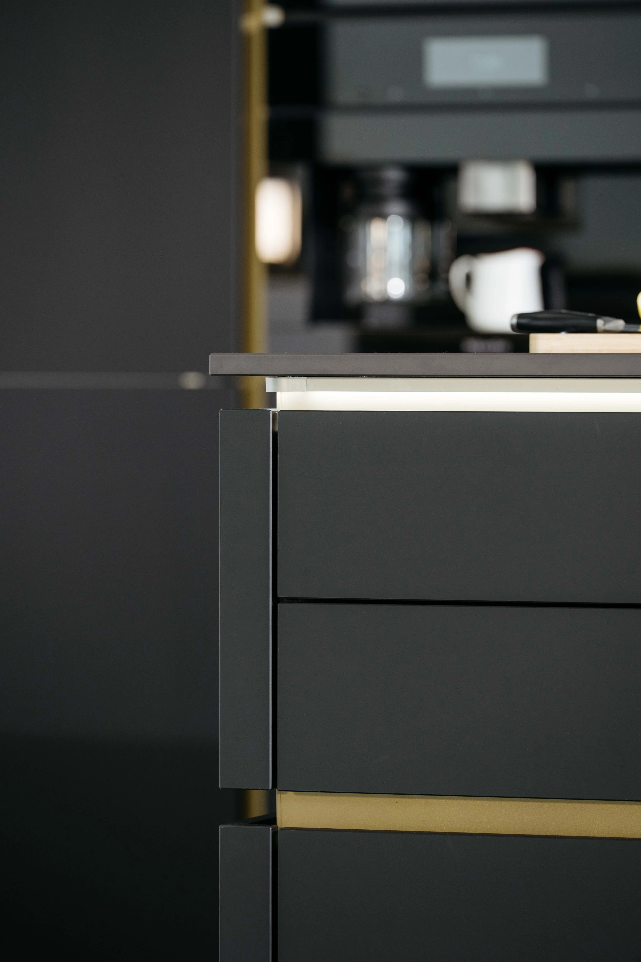 Del av ett modernt kök med svarta och guldiga skåpluckor, synliga delar av köksutrustning i bakgrunden.